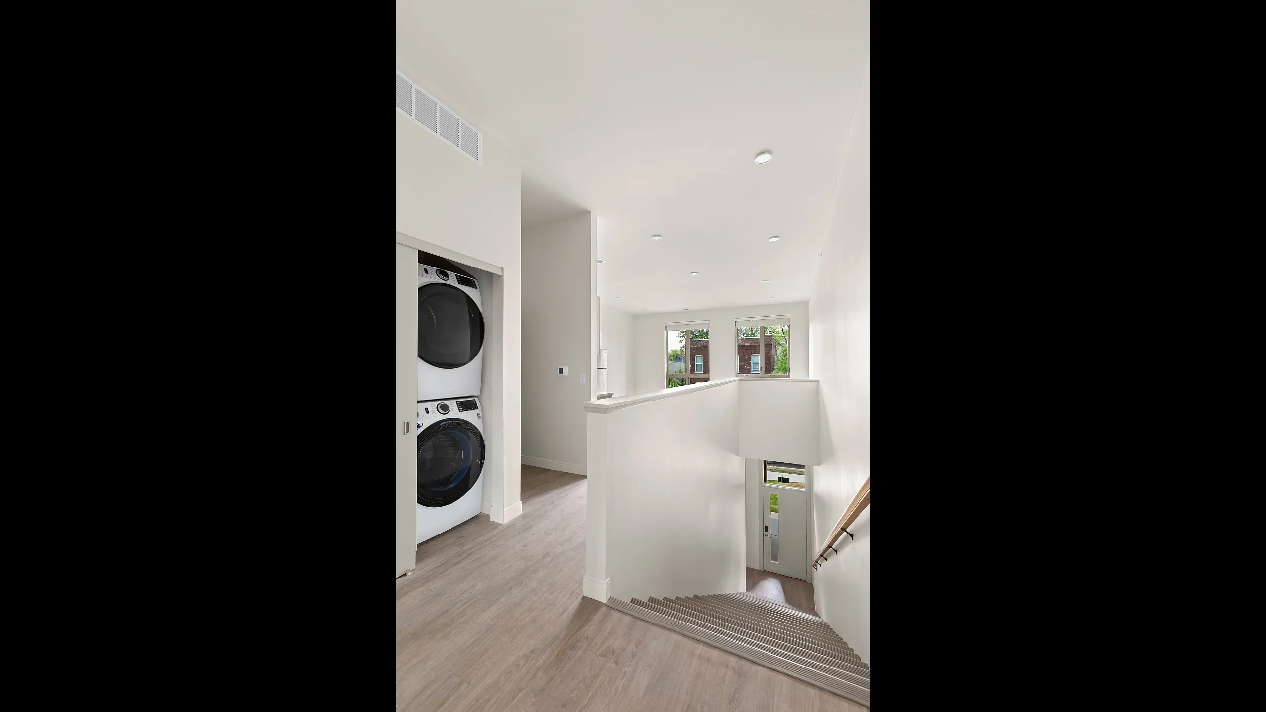 Bright modern hallway with sleek hardwood floors, featuring in-unit washer and dryer and elegant staircase leading to a sunlit entrance.