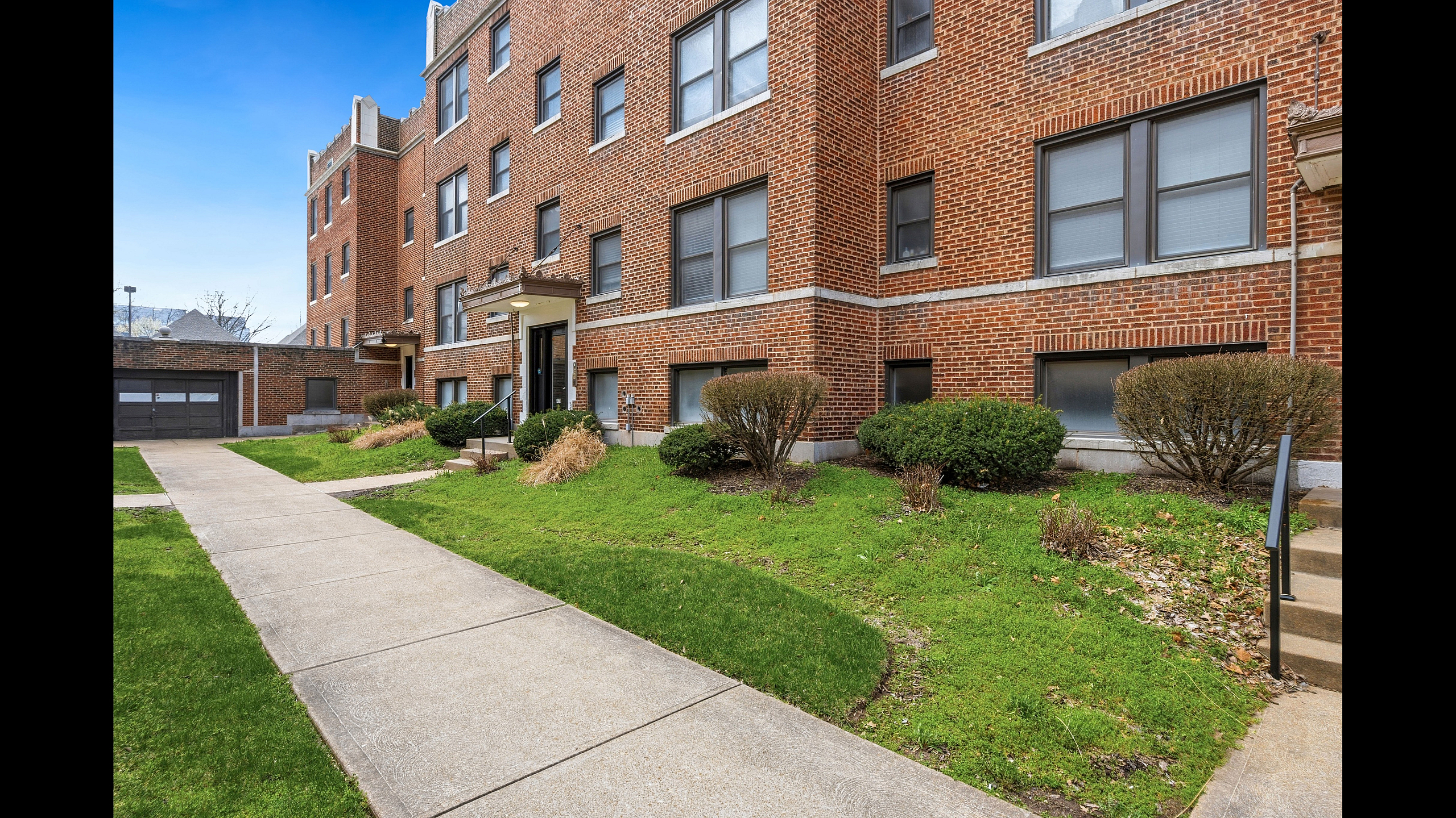Charming brick apartment building with manicured green lawns and inviting pathways, basking in bright daylight.