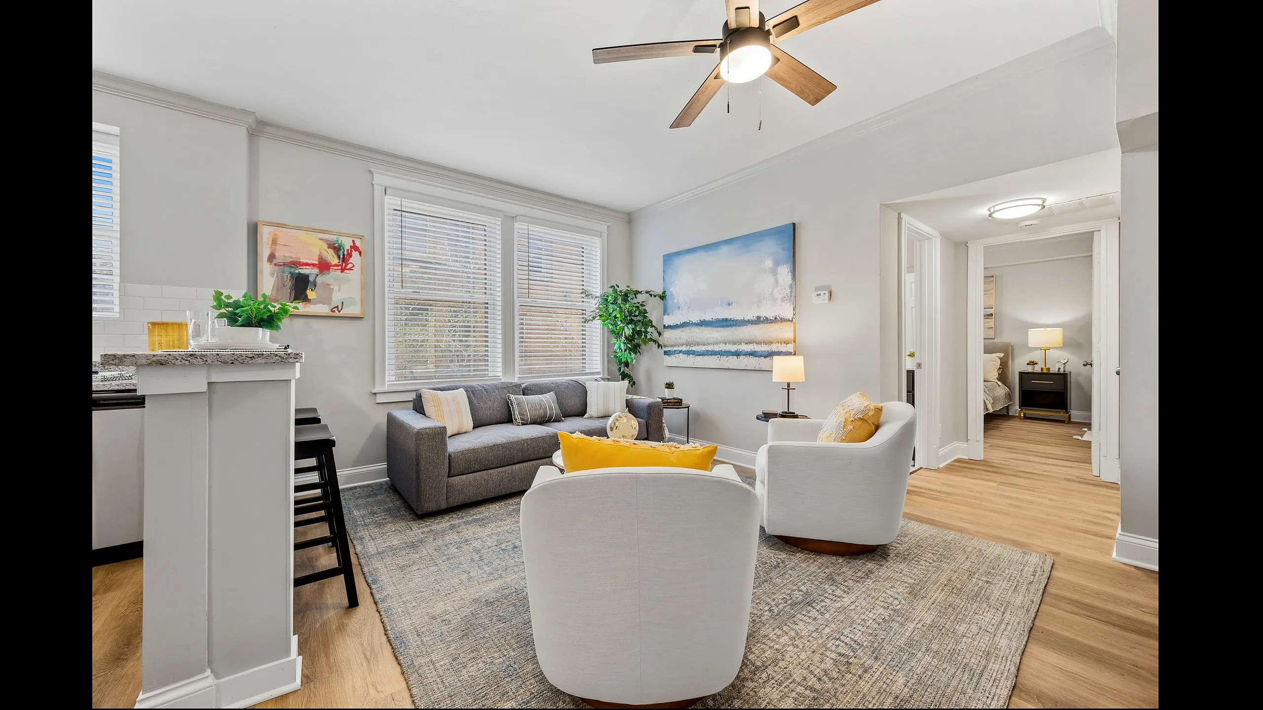 Inviting living room with modern decor, featuring a cozy gray sofa, stylish white chairs, and vibrant artwork, bathed in natural light from large windows.