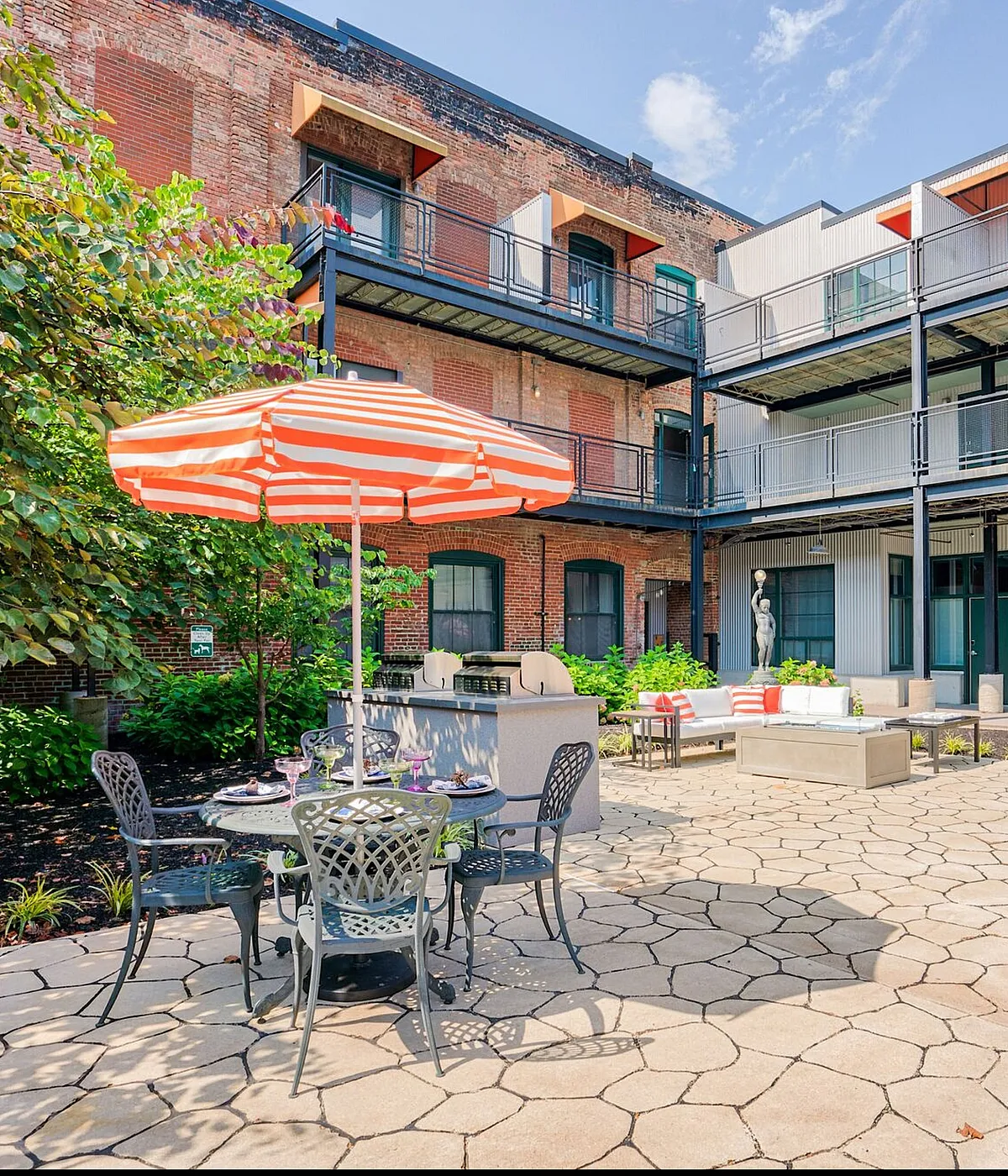 Charming courtyard of a modern apartment complex featuring stylish outdoor seating with a vibrant orange-striped umbrella, surrounded by lush greenery and a classic brick facade.