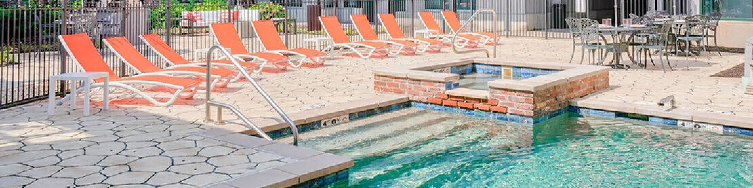 Charming apartment complex featuring a sunlit pool area with vibrant orange lounge chairs and striped umbrellas, offering an inviting space for relaxation.