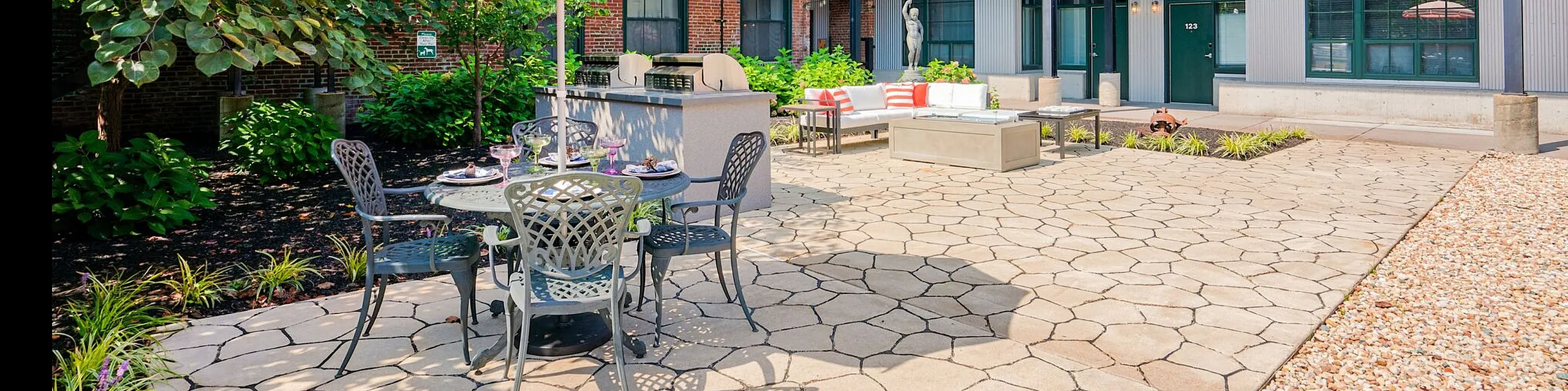 Charming courtyard patio with stylish seating and striped umbrella, surrounded by lush greenery and modern industrial architecture.