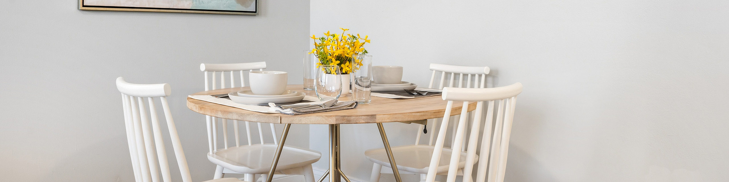 Contemporary dining nook with minimalist white chairs, round wooden table set for four, vibrant abstract wall art, and fresh yellow flowers.