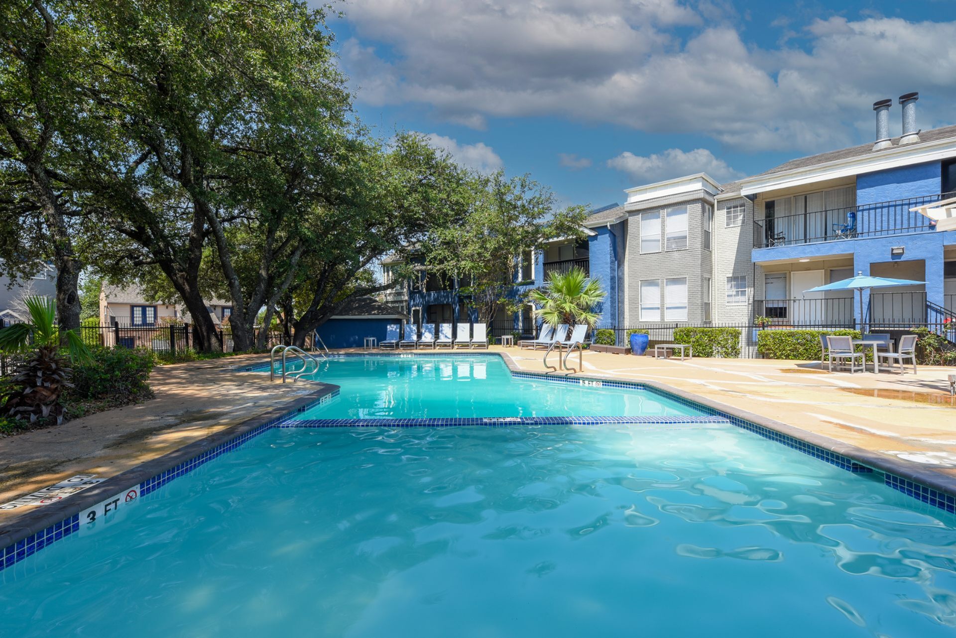 Inviting apartment complex with a pristine outdoor pool, surrounded by lush trees and stylish loungers under a bright blue sky.