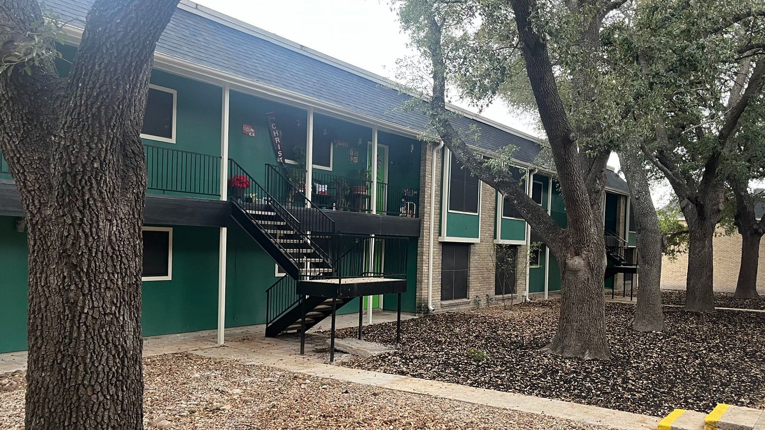 Charming two-story apartment building with green accents, nestled among mature trees offering a serene atmosphere.