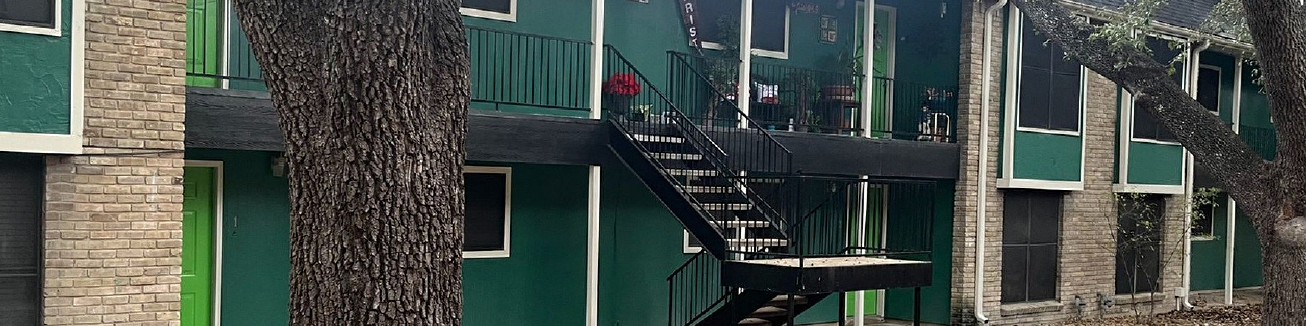 Charming two-story apartment building with vibrant green accents and a welcoming outdoor staircase, nestled among mature trees.