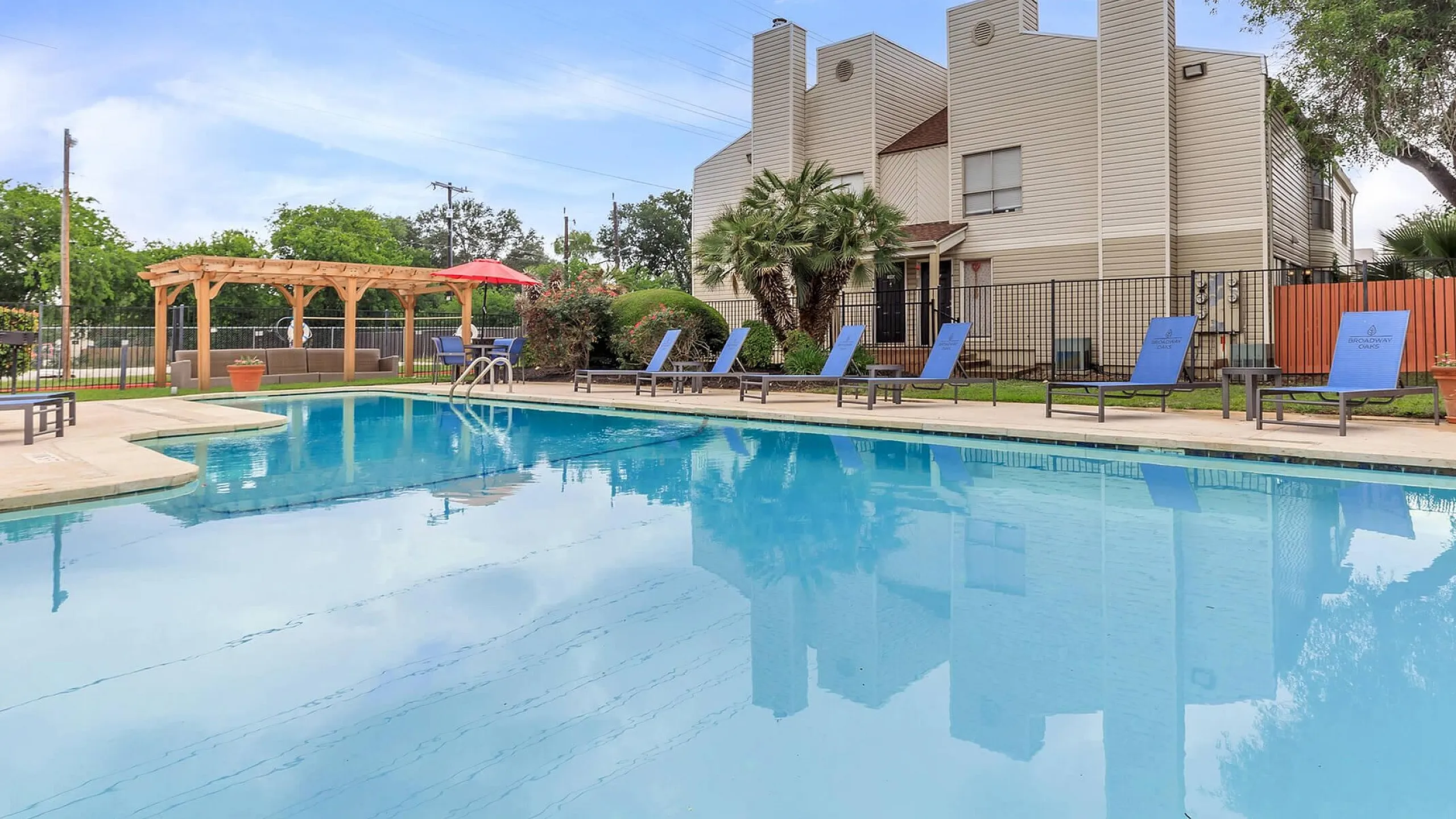 Inviting apartment complex with a sparkling pool, surrounded by lounge chairs and a charming pergola, offering a perfect relaxation spot in a serene setting.