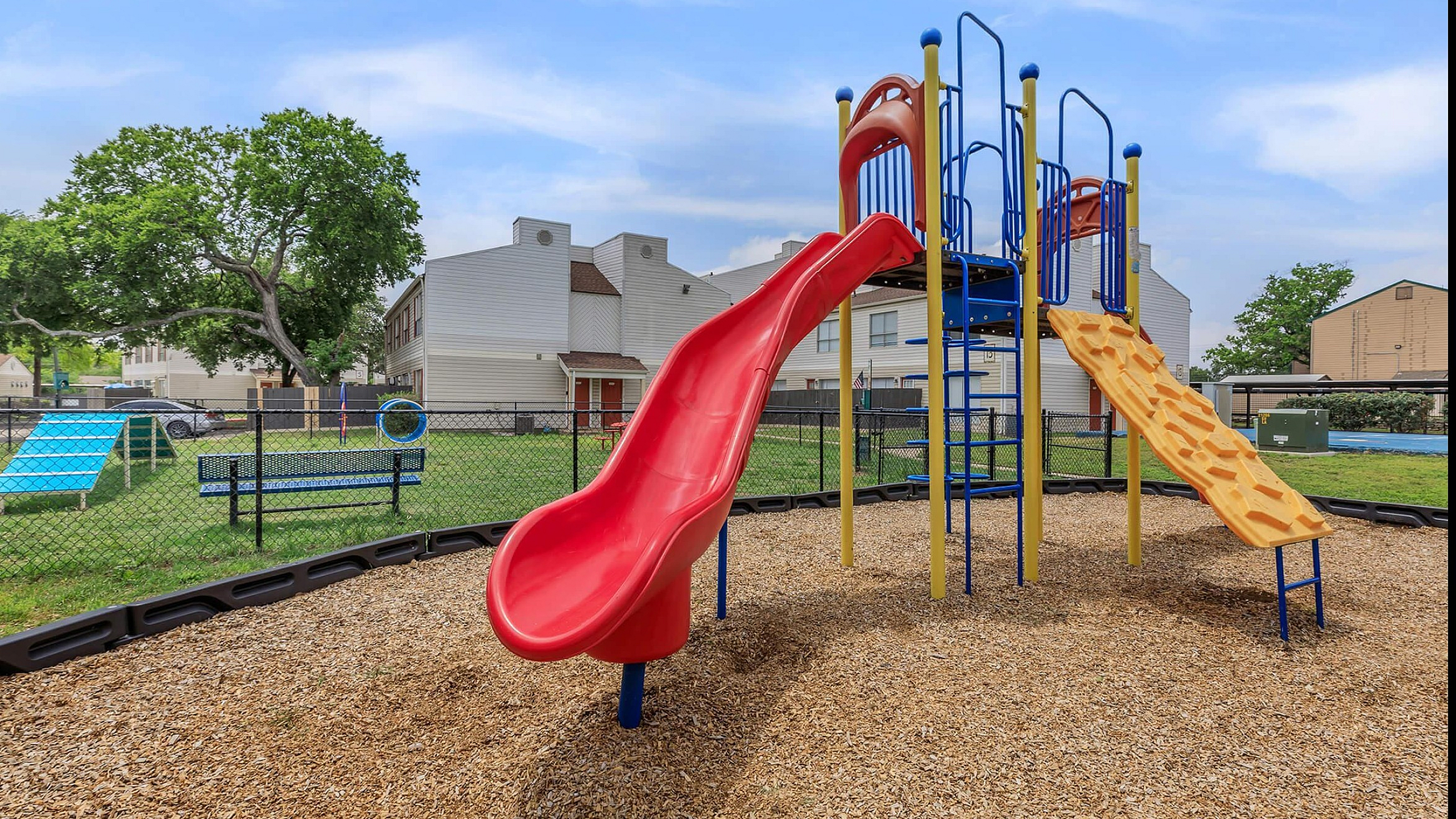 Vibrant playground with a towering red slide, climbing structure, and picnic benches, set in a sunny residential community.