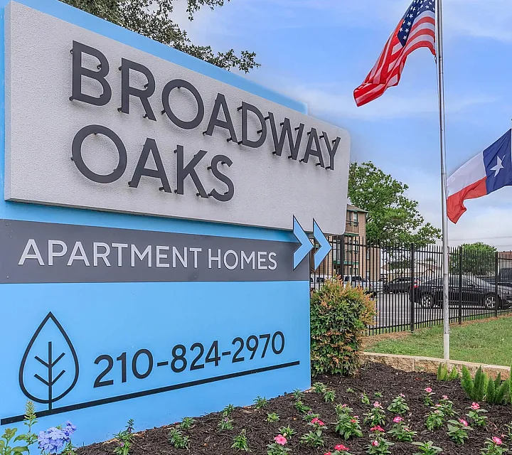 Inviting entrance to Broadway Oaks Apartment Homes with vibrant landscaping and flags, showcasing a welcoming community atmosphere.