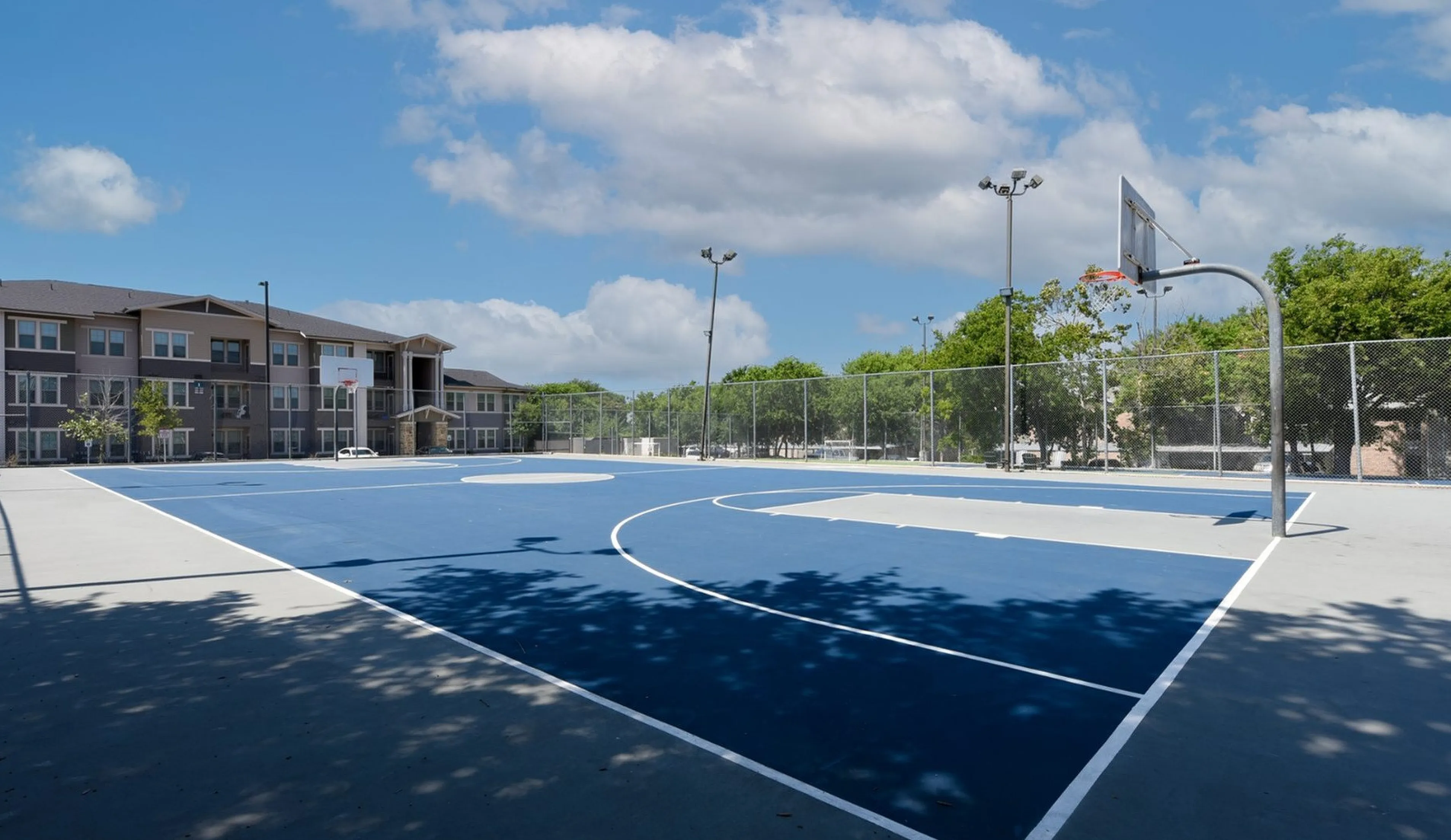 Modern apartment complex with scenic outdoor basketball court under a clear blue sky, surrounded by lush greenery, offering an active lifestyle.
