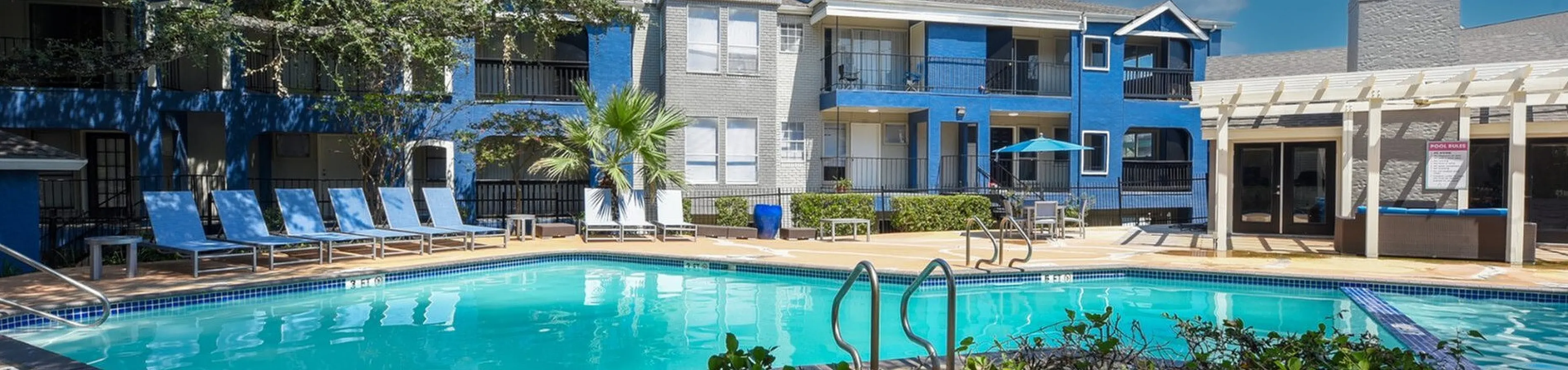 Charming apartment complex with a refreshing pool, elegant sun loungers, and lush greenery under a vibrant blue sky.