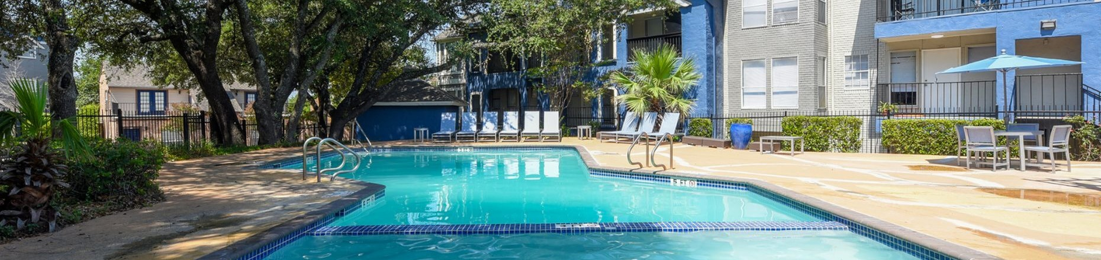 Inviting apartment complex featuring a sparkling outdoor swimming pool surrounded by lush greenery and modern architecture.
