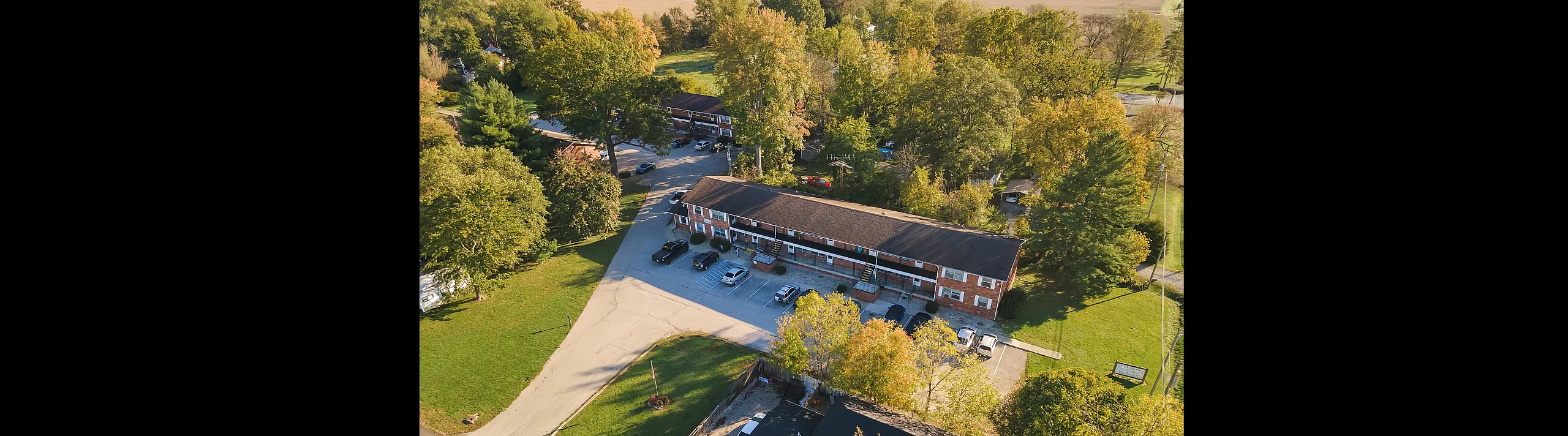 Charming aerial view of a brick apartment complex nestled among lush green trees, offering ample parking and serene surroundings.