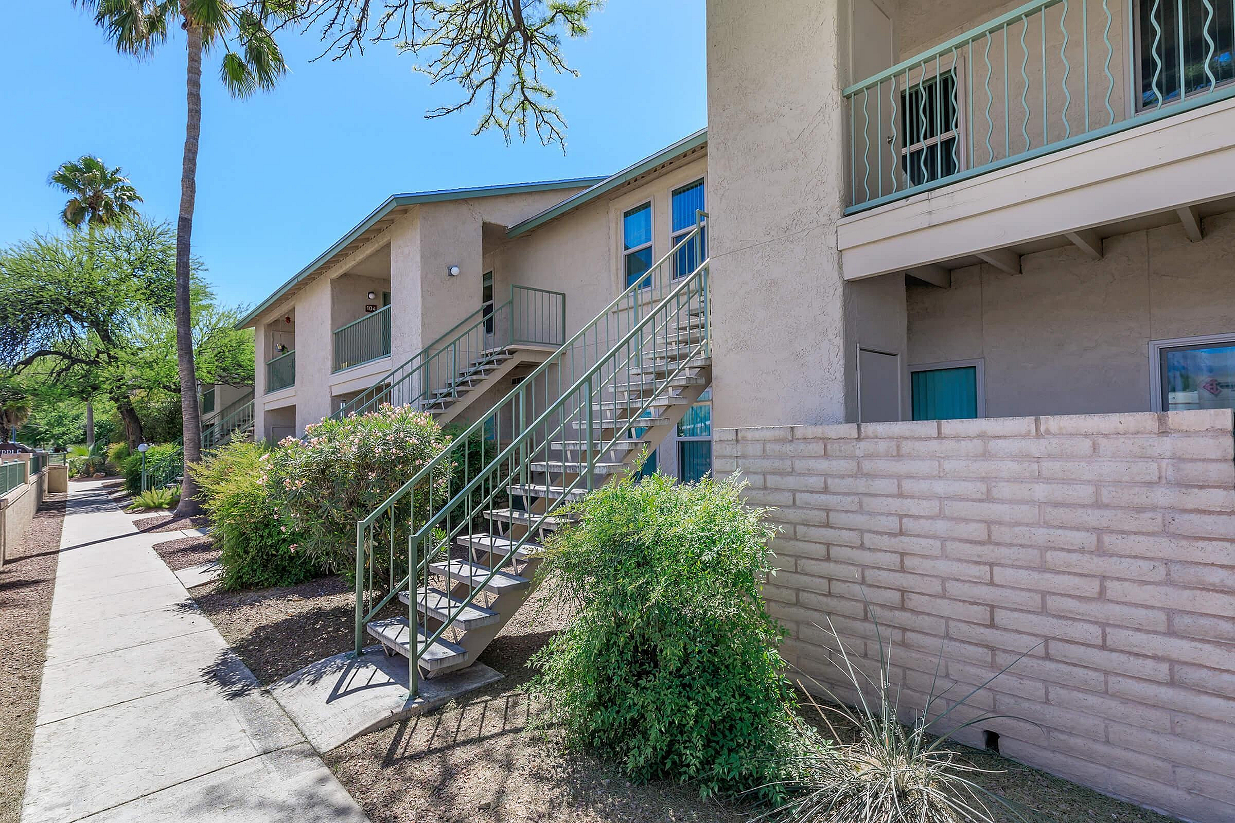 Charming apartment exterior with stylish staircases and lush greenery, framed by clear blue skies and palm trees.