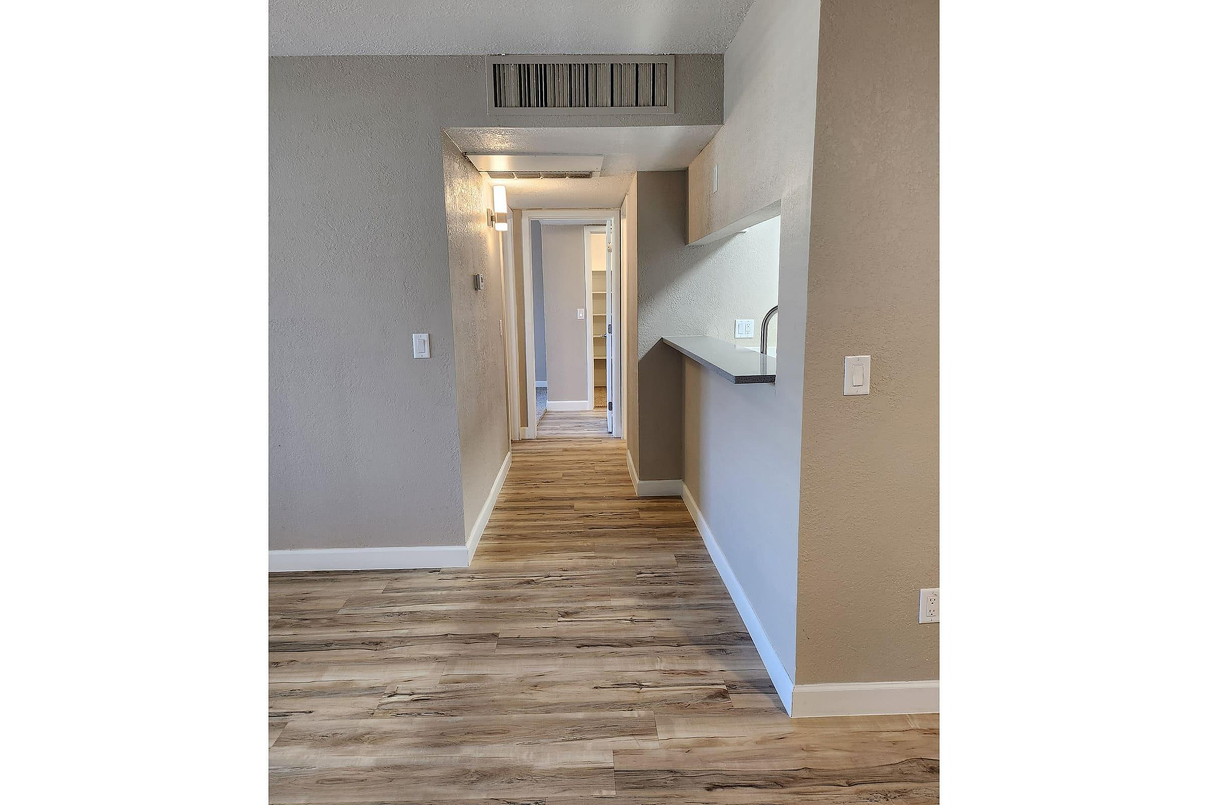 Bright apartment hallway with elegant wood flooring and modern lighting, leading to a cozy room with built-in shelving.