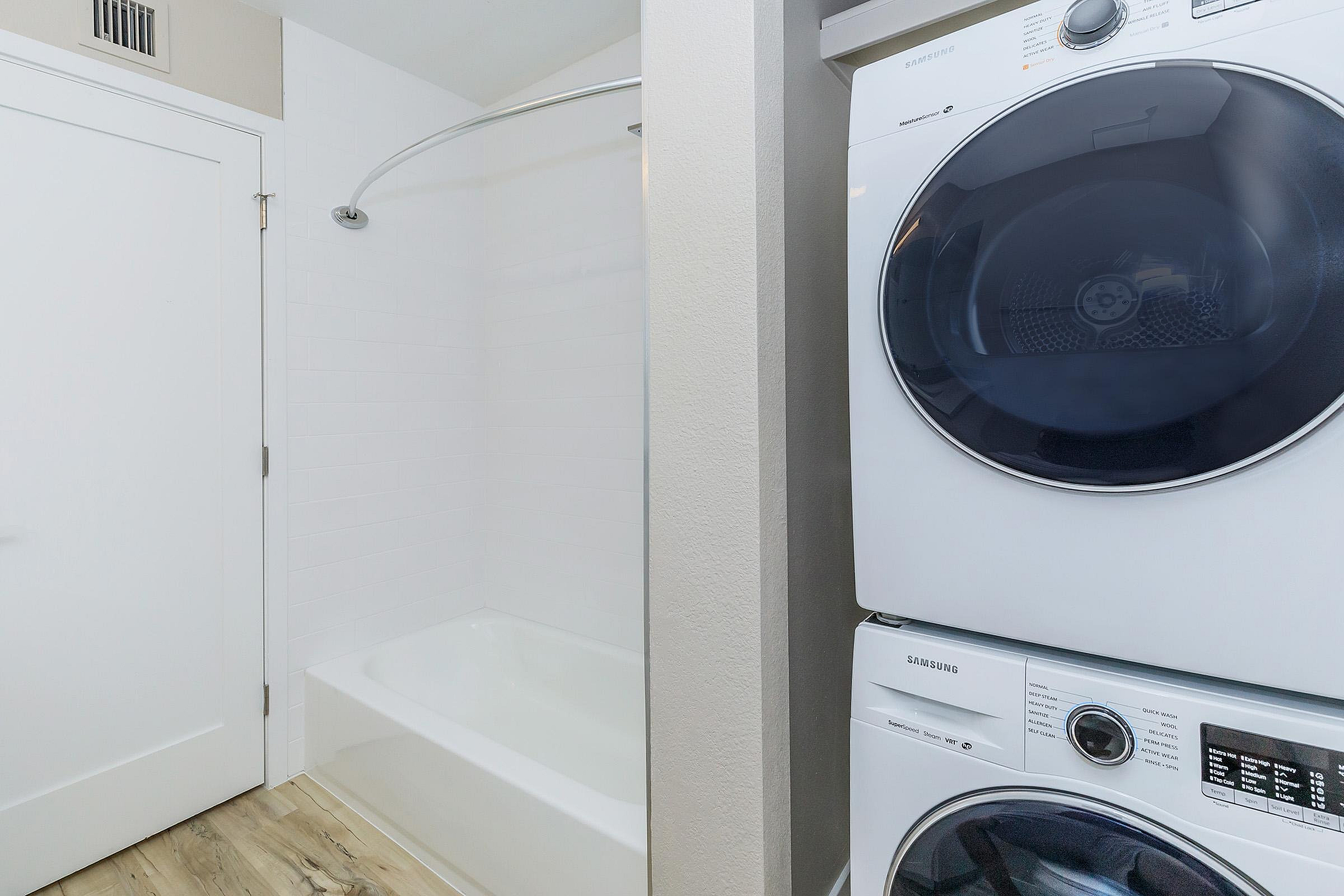 Modern bathroom with a sleek shower and in-unit stacked Samsung washer and dryer, featuring elegant white tile and warm wood flooring.