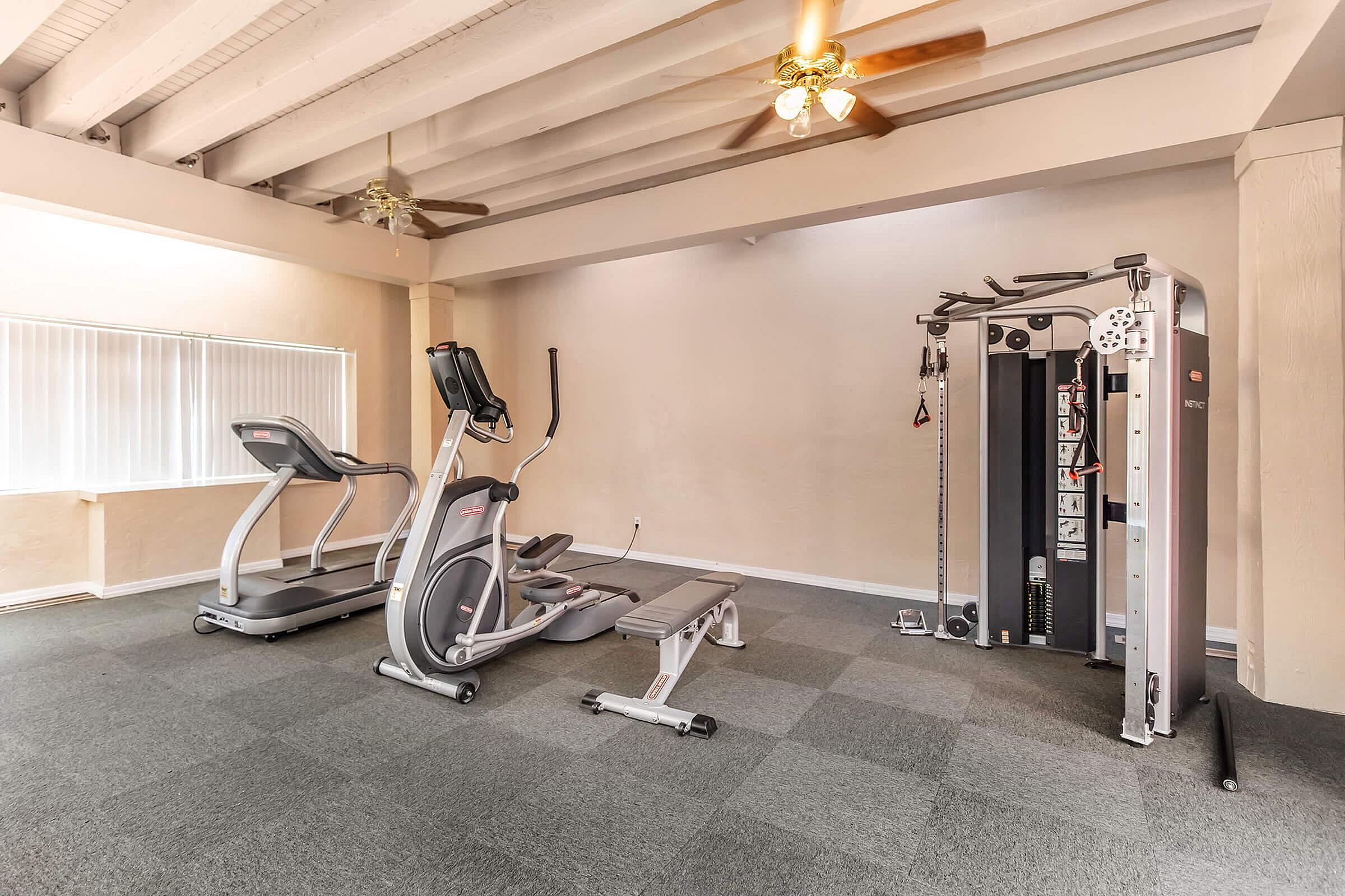 Bright and spacious home gym featuring modern fitness equipment, including an elliptical, treadmill, and multifunctional weight machine under stylish ceiling fans.