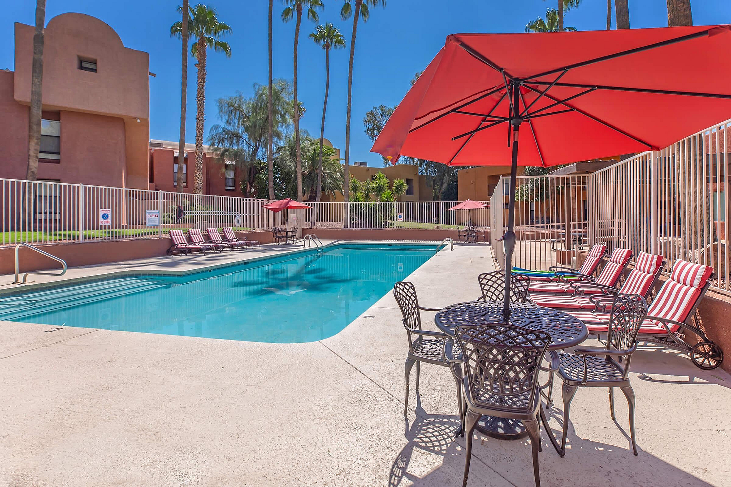 Inviting outdoor pool area with striped lounge chairs, dining table, and vibrant red umbrellas surrounded by lush palm trees.