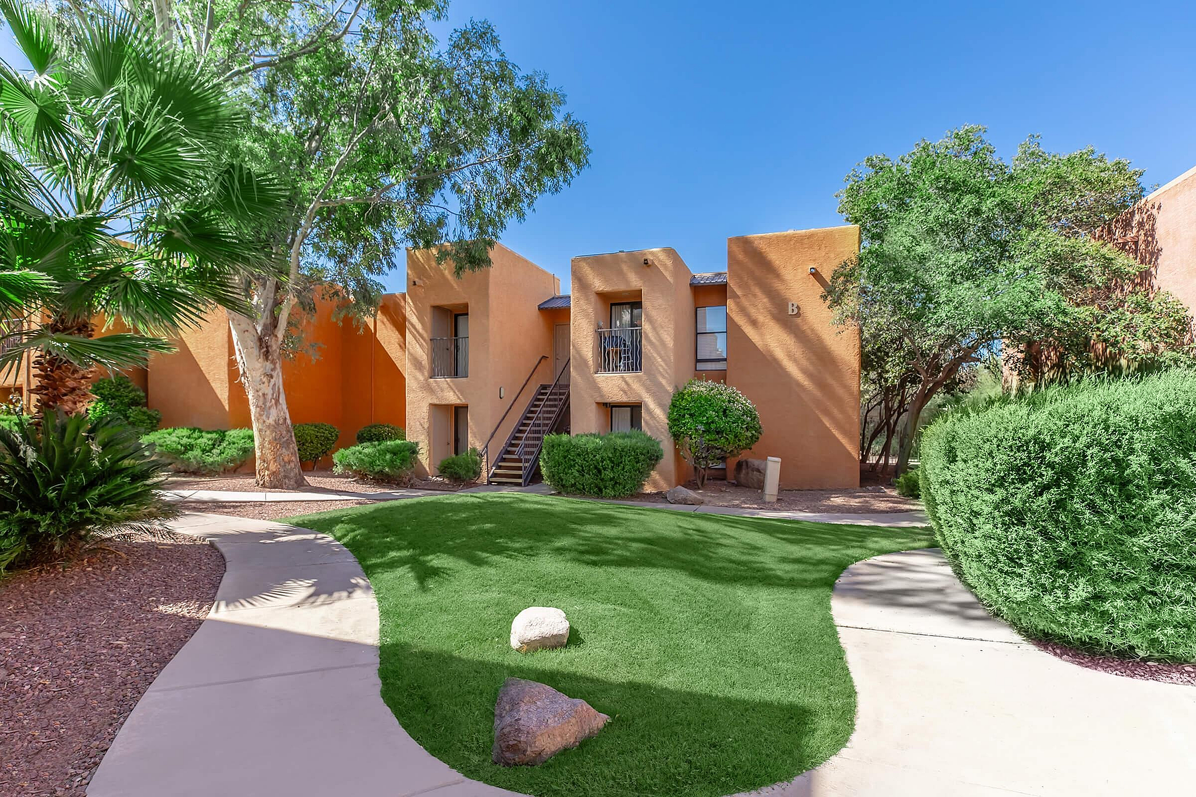 Charming desert-style apartment complex with vibrant greenery, featuring a sunny walkway leading to inviting staircases.