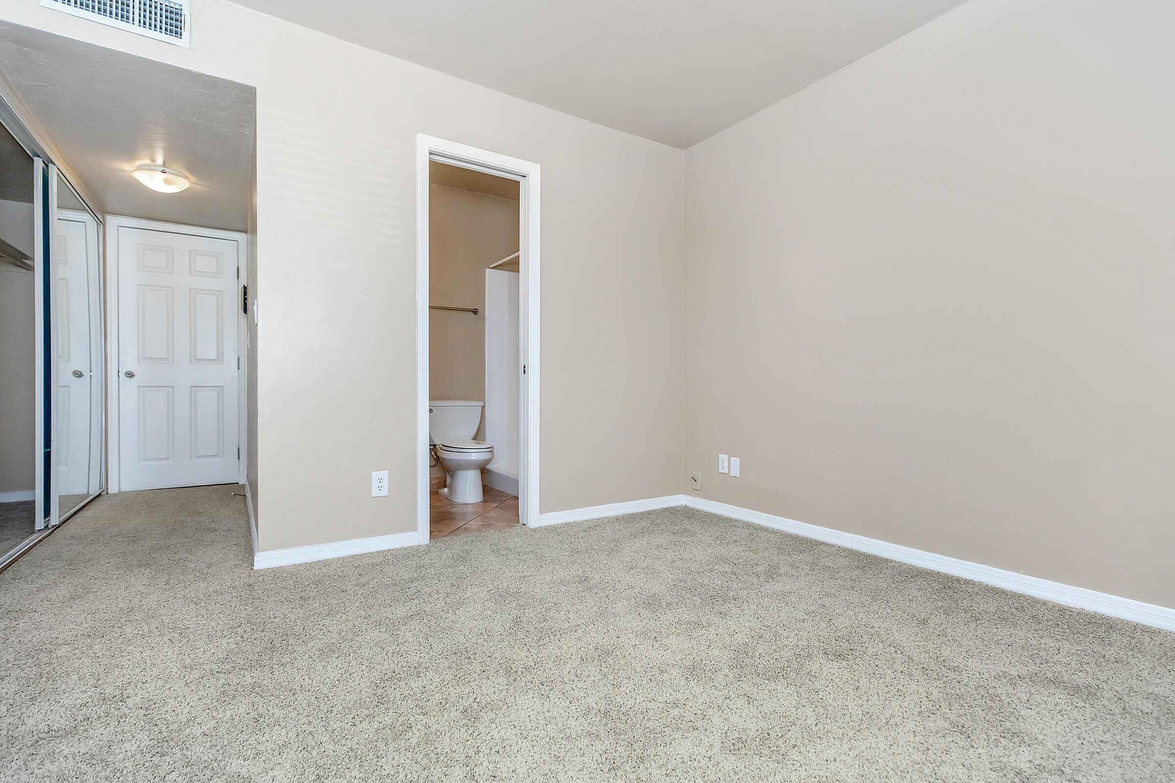 Bright and airy bedroom with plush carpeting and ensuite bathroom, featuring mirrored closet doors for a spacious feel.