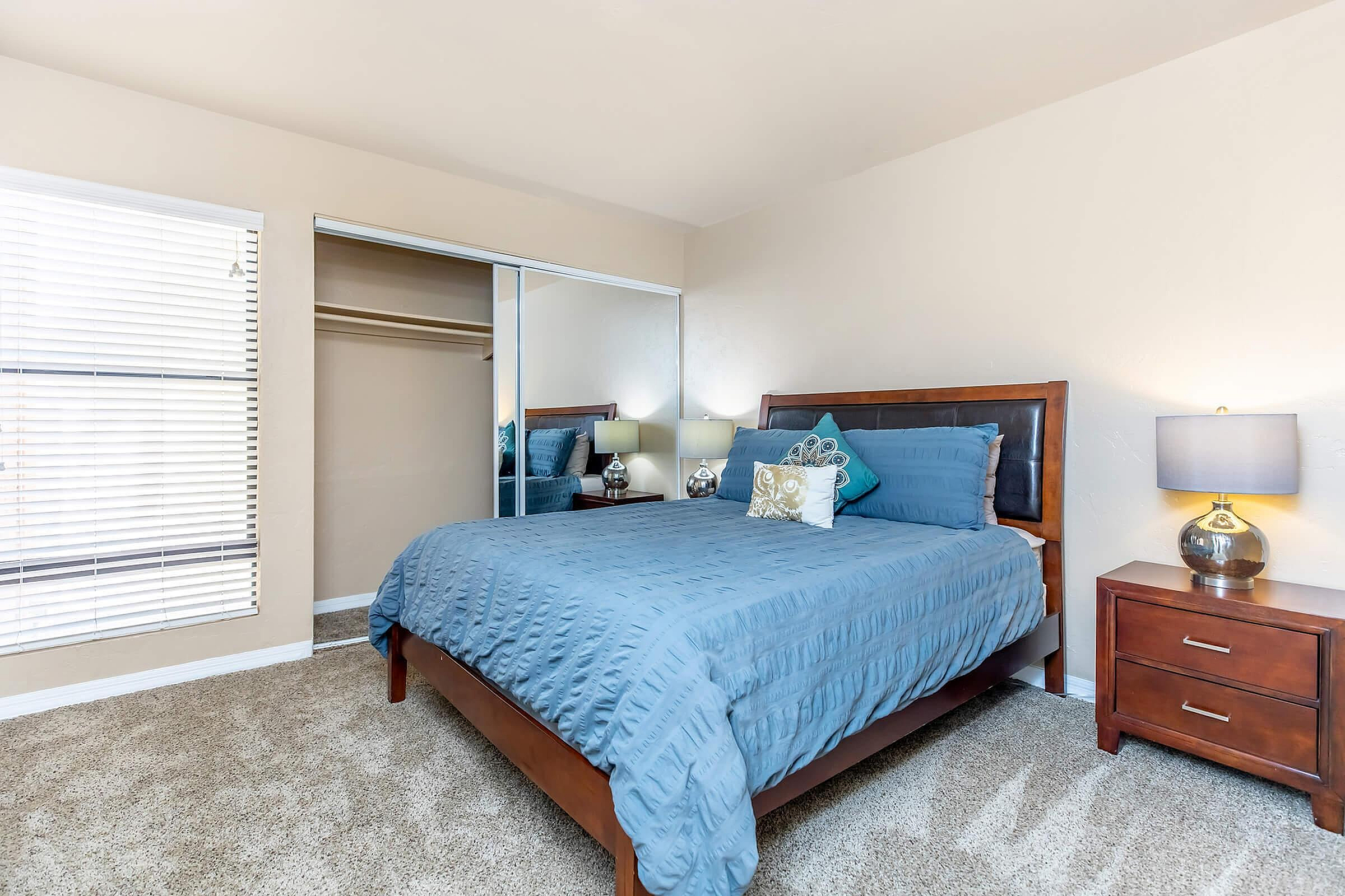 Inviting bedroom with plush carpeting, elegant wooden bed frame, mirrored closet, and stylish bedside lamps.