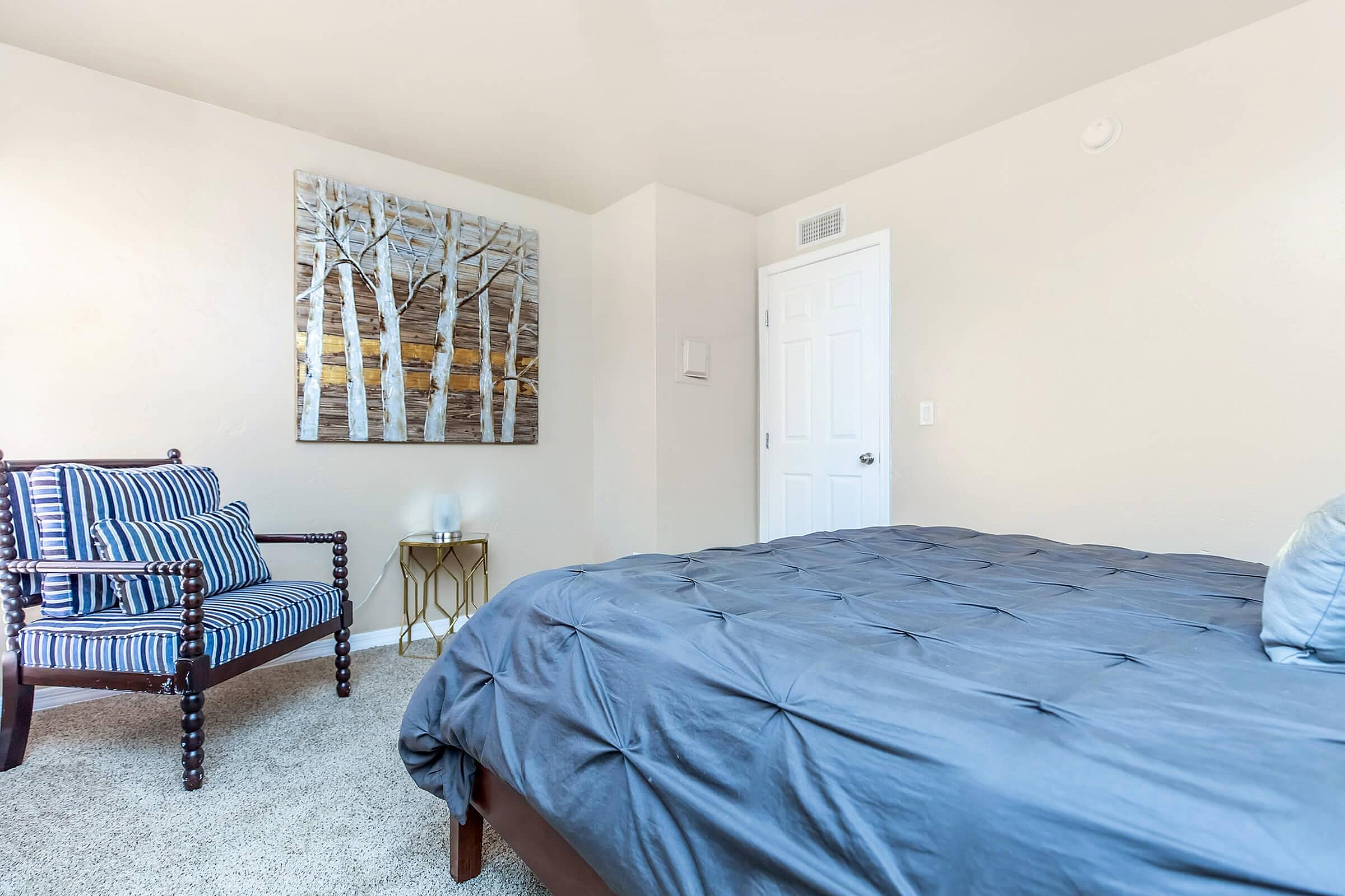 Bright and cozy bedroom featuring a comfortable bed with a stylish blue comforter, an elegant striped chair, and modern artwork on a light-colored wall.