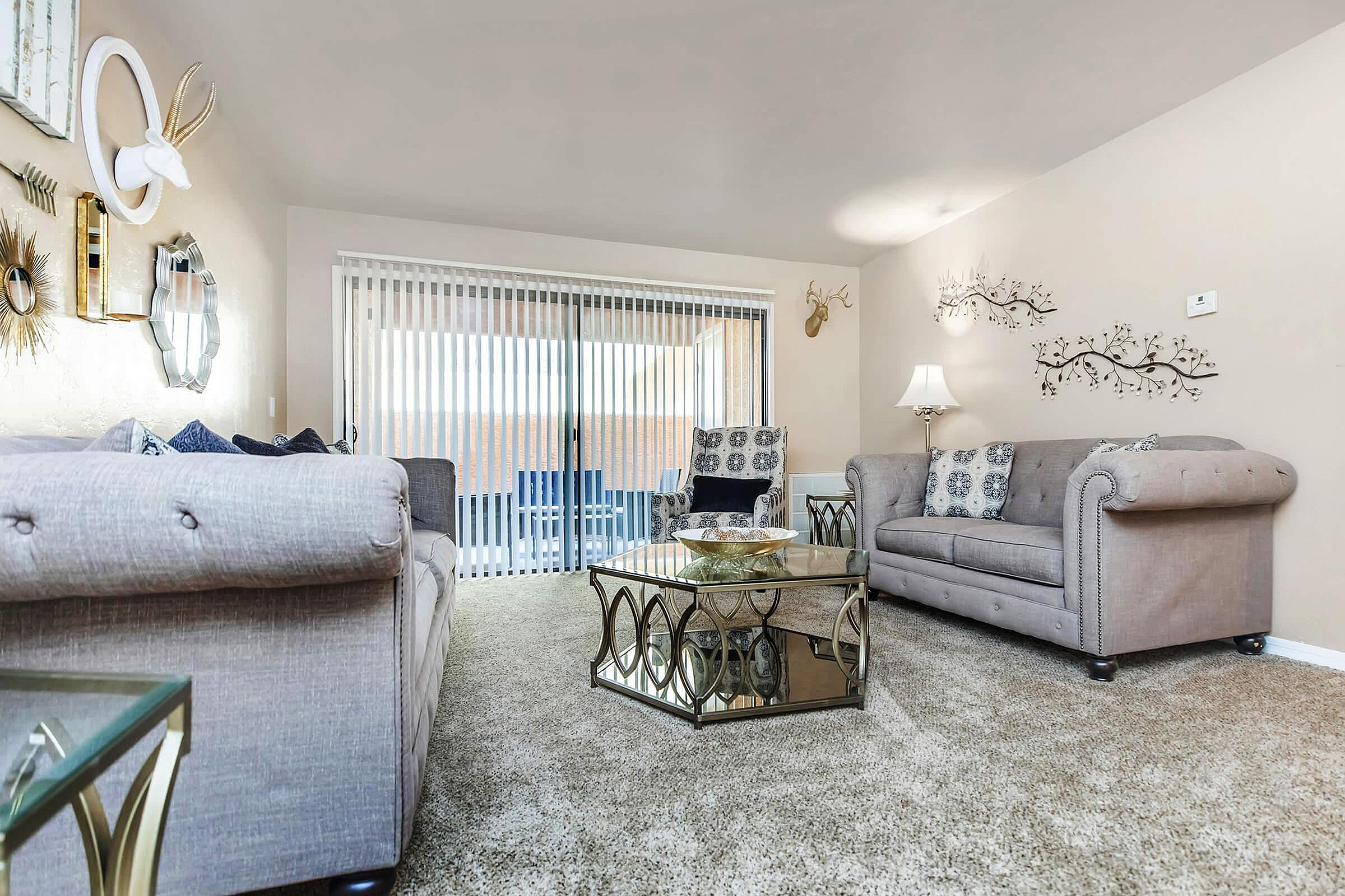 Inviting living room with plush beige sofas, elegant decor, and a glass coffee table; abundant natural light streaming through large sliding glass doors with vertical blinds.