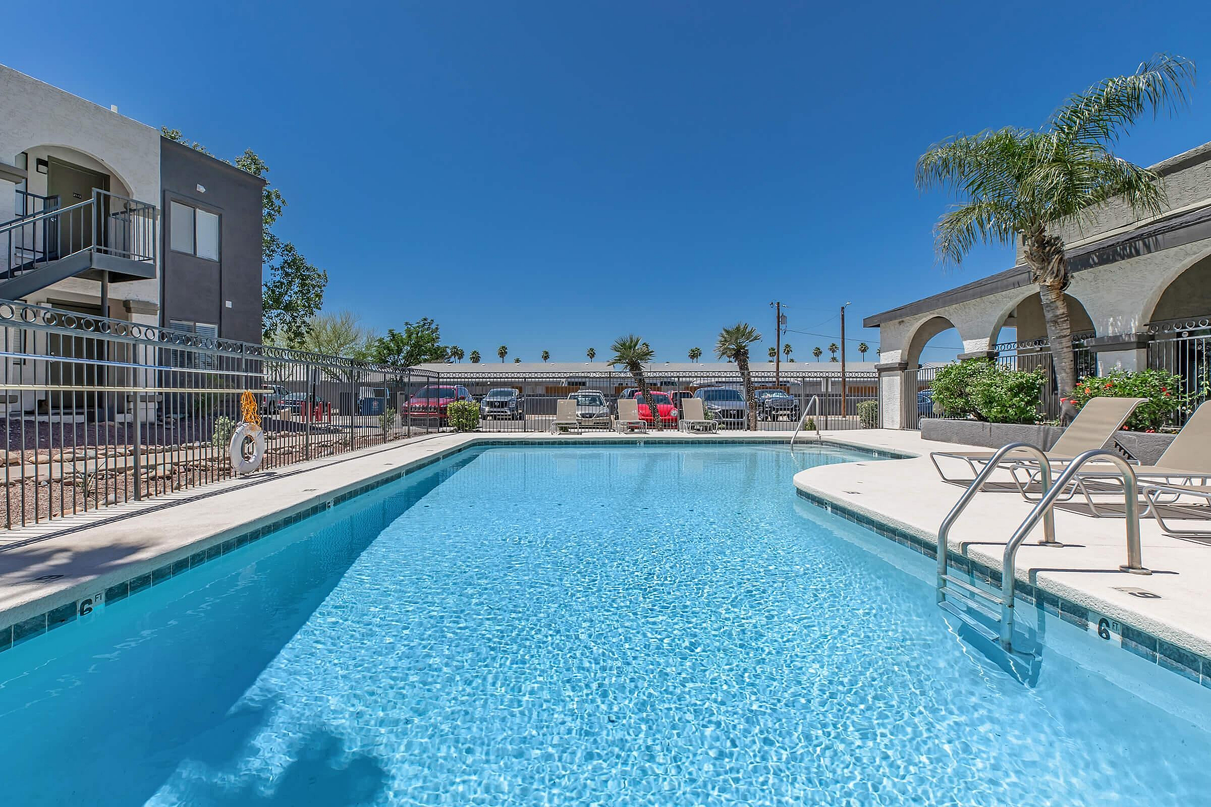 Inviting apartment pool with crystal-clear water, surrounded by lush palm trees and comfortable lounge chairs, under a clear blue sky.