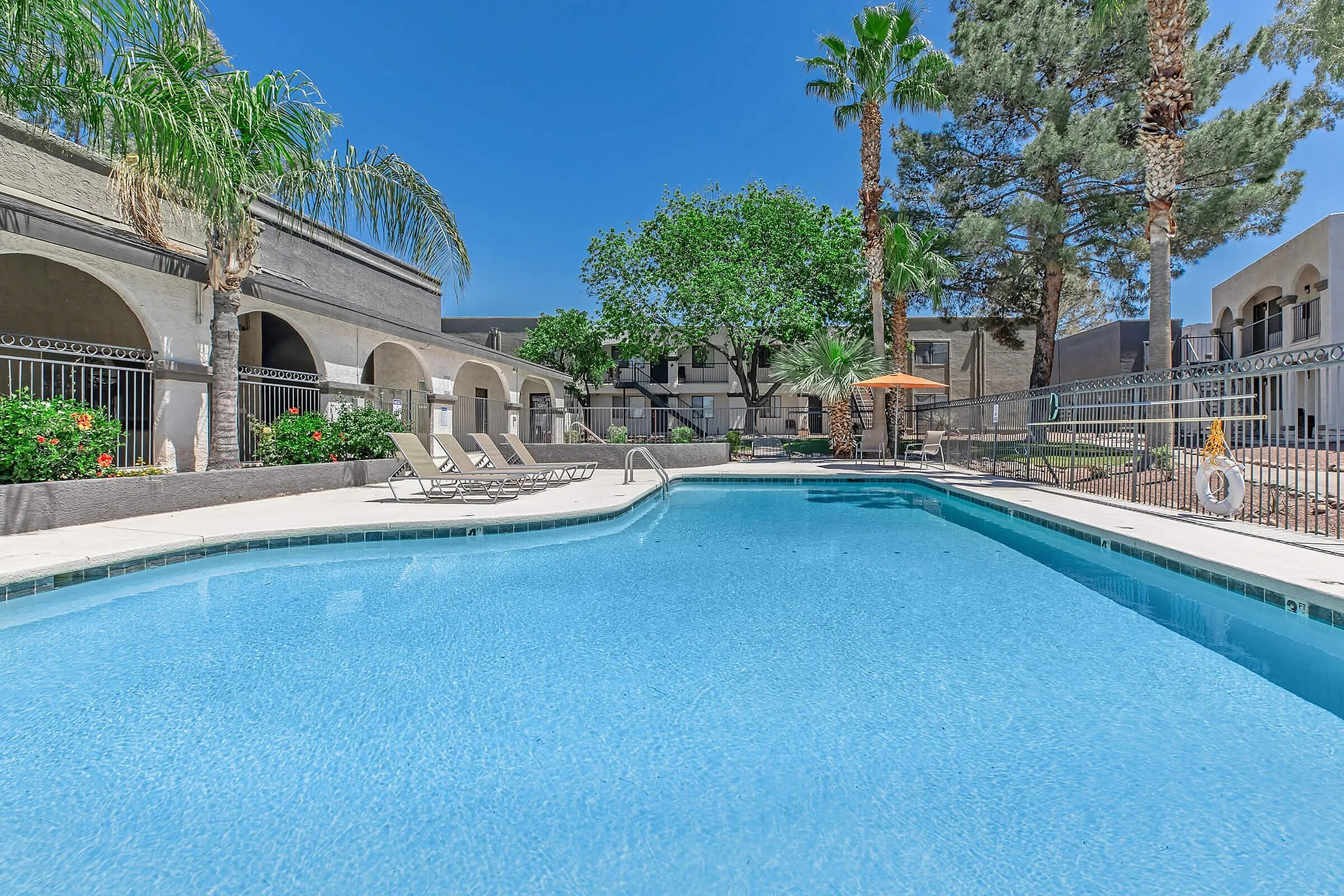 Inviting apartment complex pool surrounded by lush greenery and elegant arches, offering a serene oasis with comfortable lounge chairs under a bright blue sky.