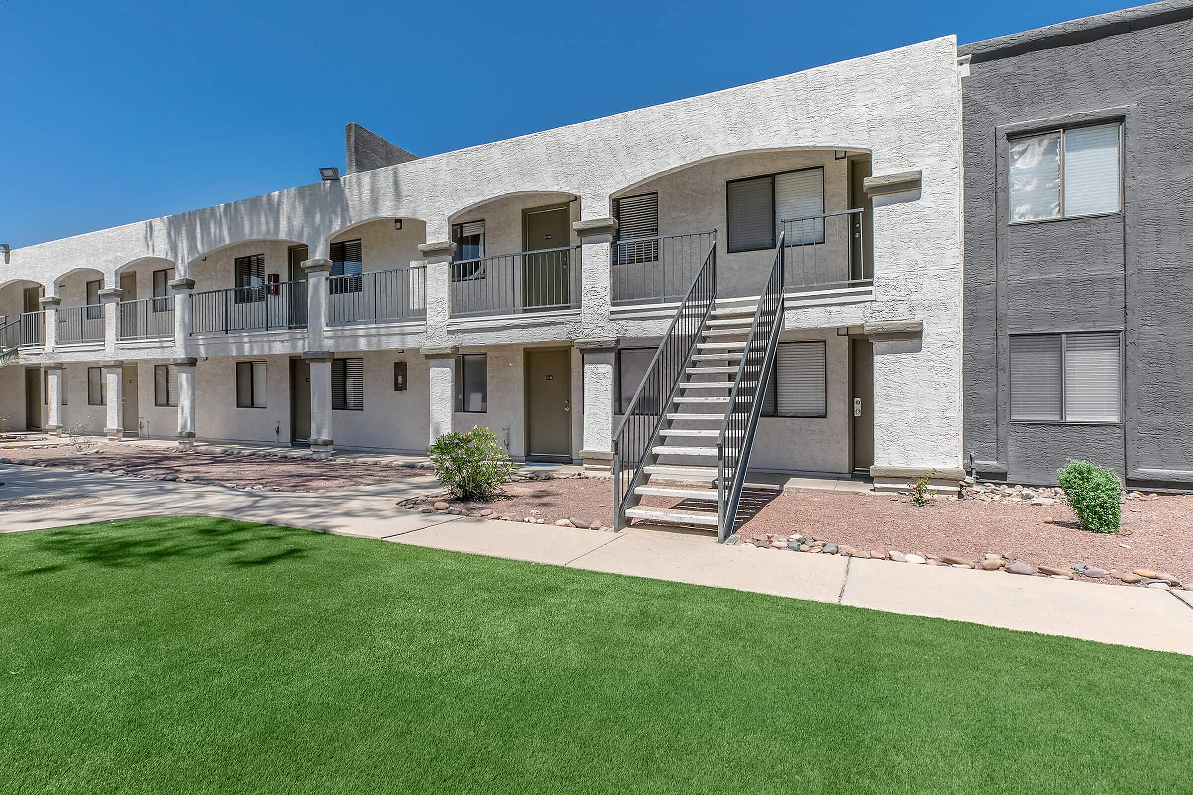 Modern apartment complex with a clean, white exterior and lush green lawn, featuring inviting balconies and a central staircase under a clear blue sky.