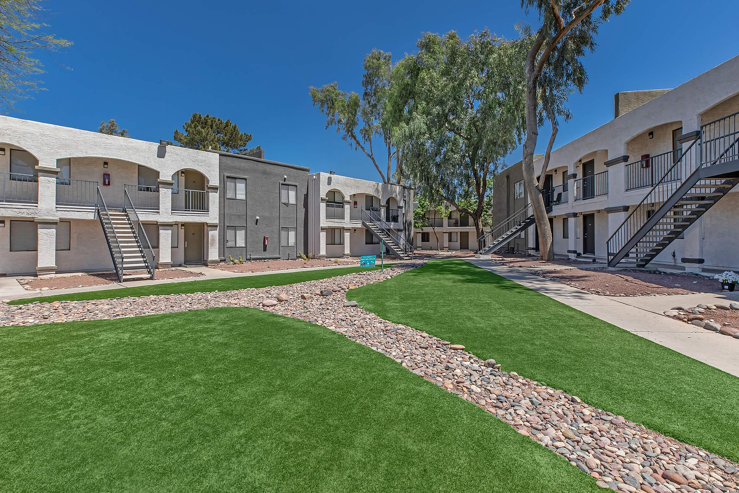 Charming apartment complex courtyard featuring lush green lawn, beautiful stone pathways, and modern architecture under a clear blue sky.