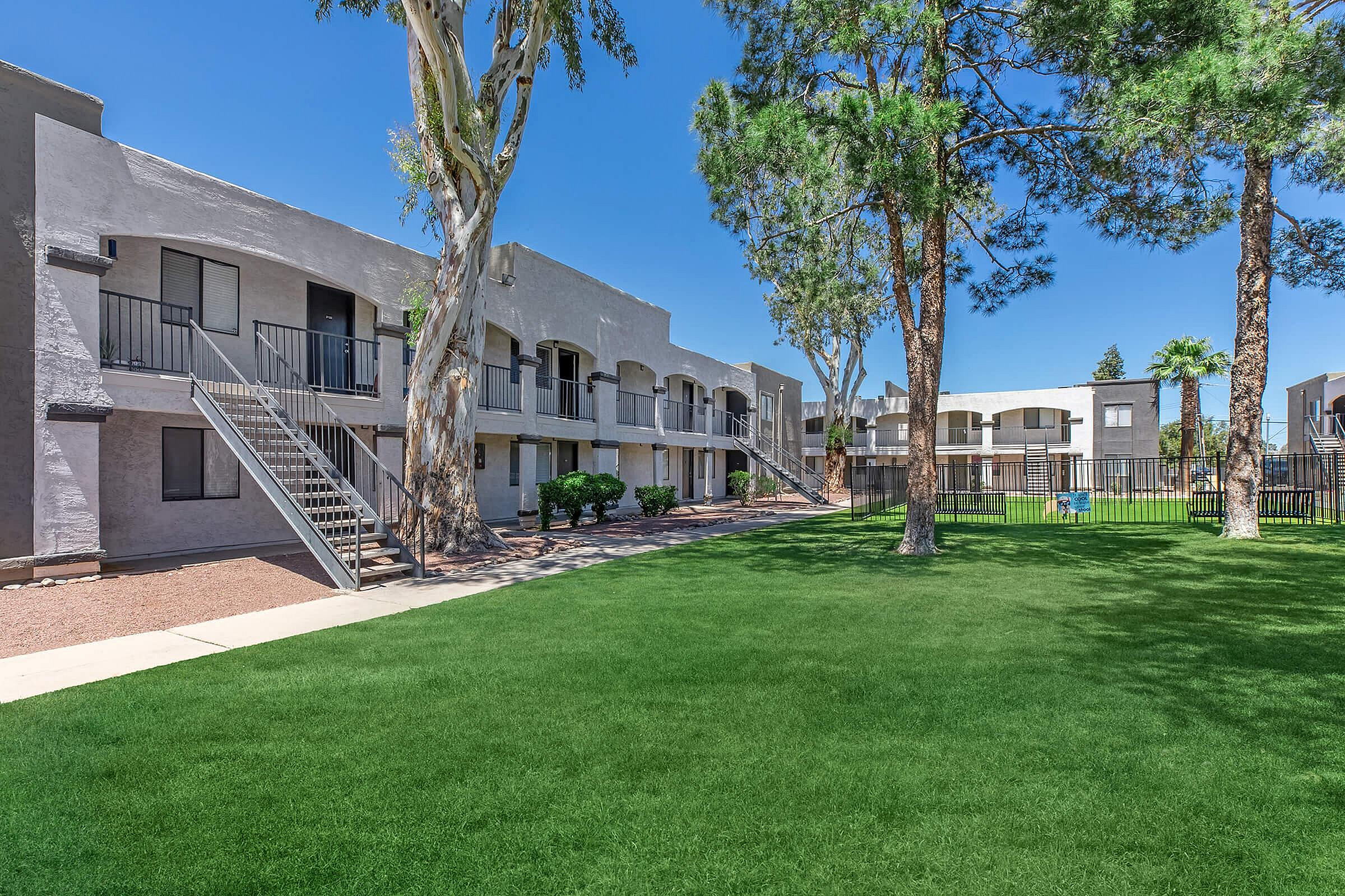 Charming apartment complex with lush green lawns, modern balconies, and vibrant trees under a clear blue sky.