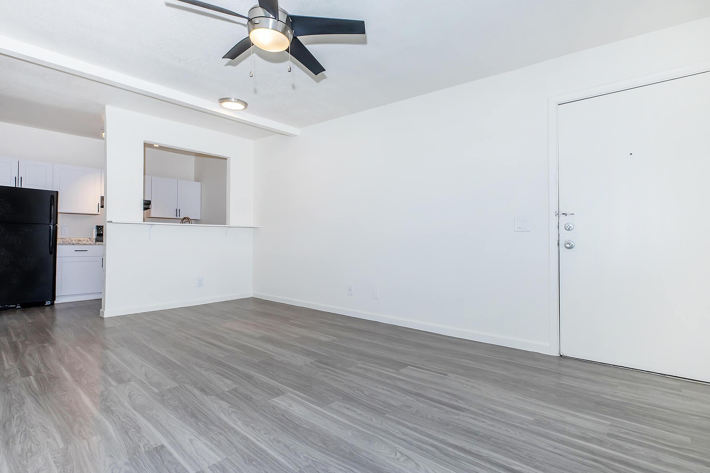 Spacious open-plan living area featuring sleek hardwood floors and modern ceiling fan, with a view into a contemporary kitchen.