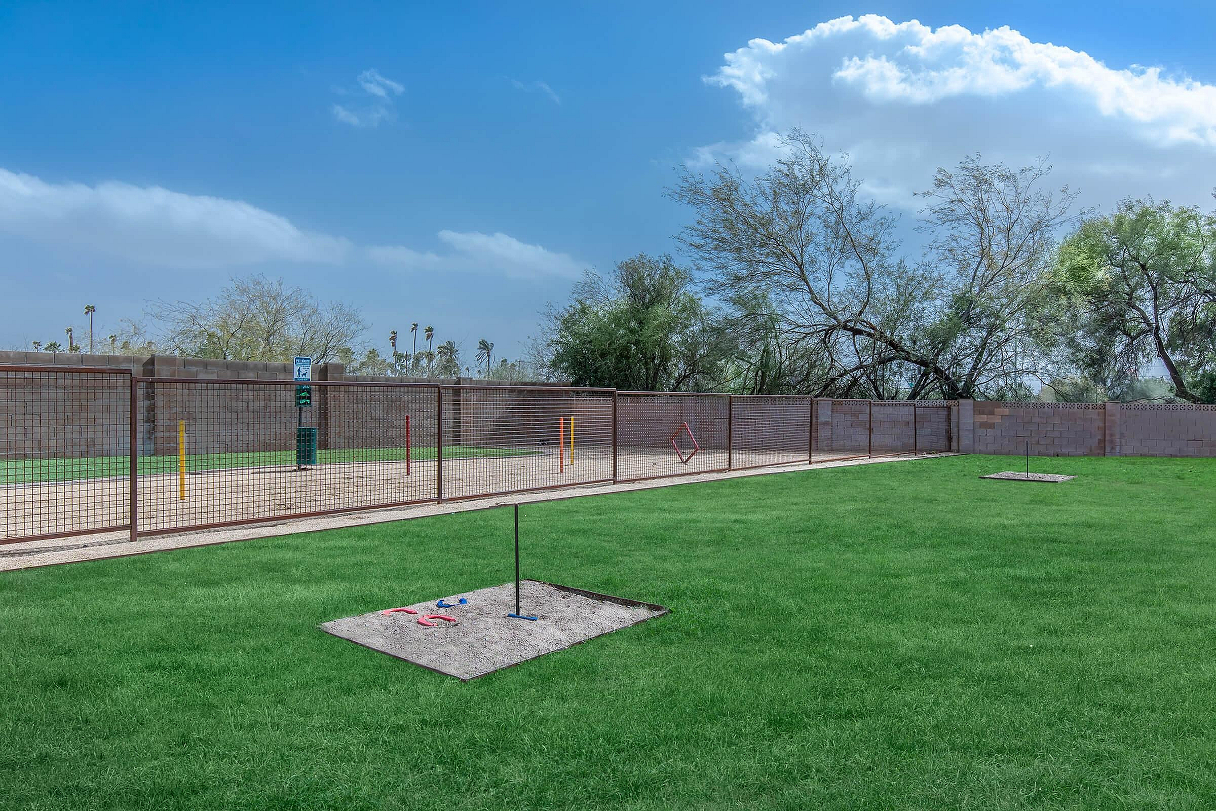 Scenic dog park with agility equipment and lush greenery, set against a backdrop of clear blue skies and trees.