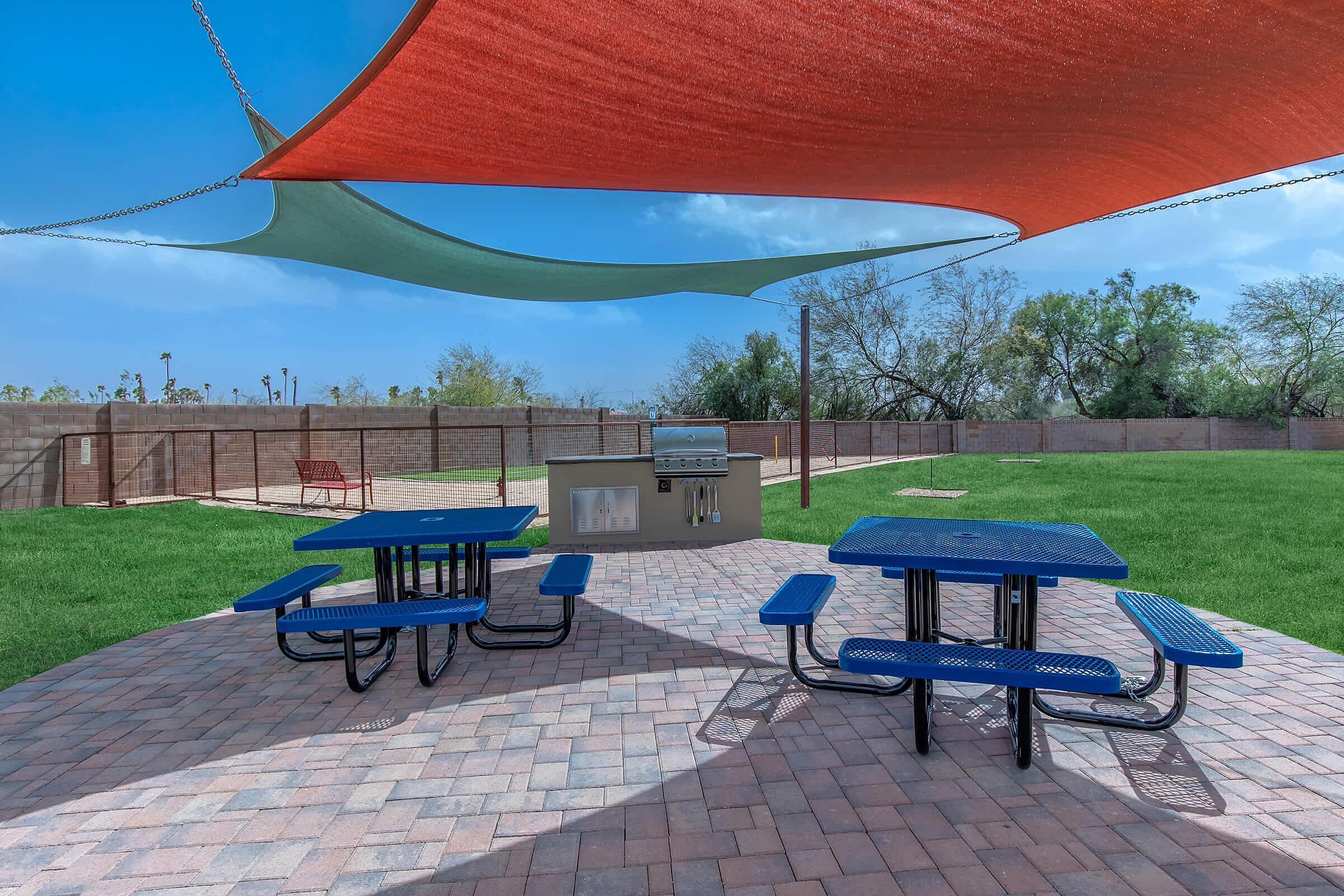 Inviting outdoor picnic area featuring vibrant blue tables, brick patio, and modern grill under colorful shade sails, surrounded by lush greenery.