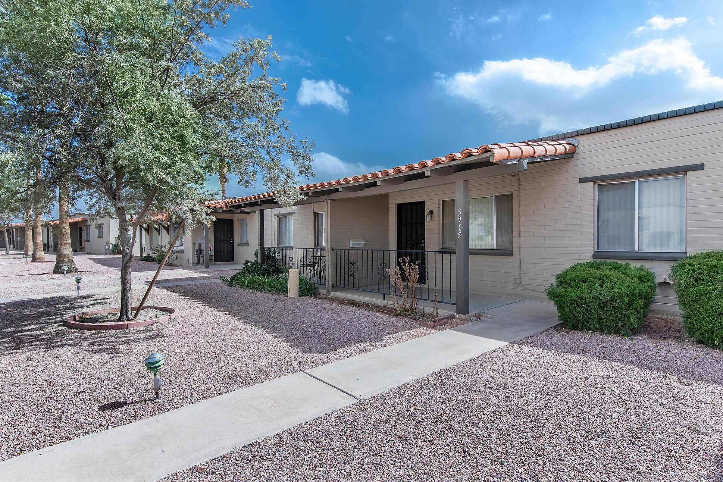 Charming single-story apartments with terracotta roof tiles, nestled among mature trees and landscaped paths, offering a serene desert ambiance.