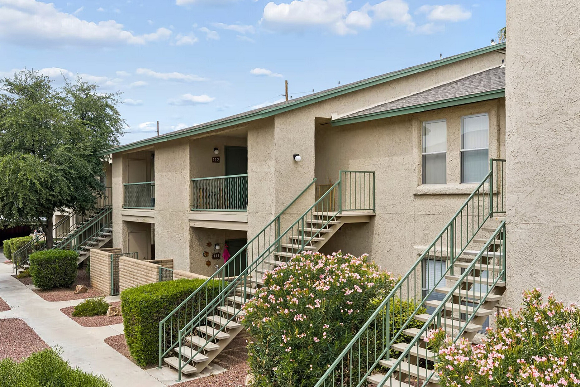 Charming two-story apartment building with lush landscaping, featuring external staircases and inviting balconies under a bright blue sky.