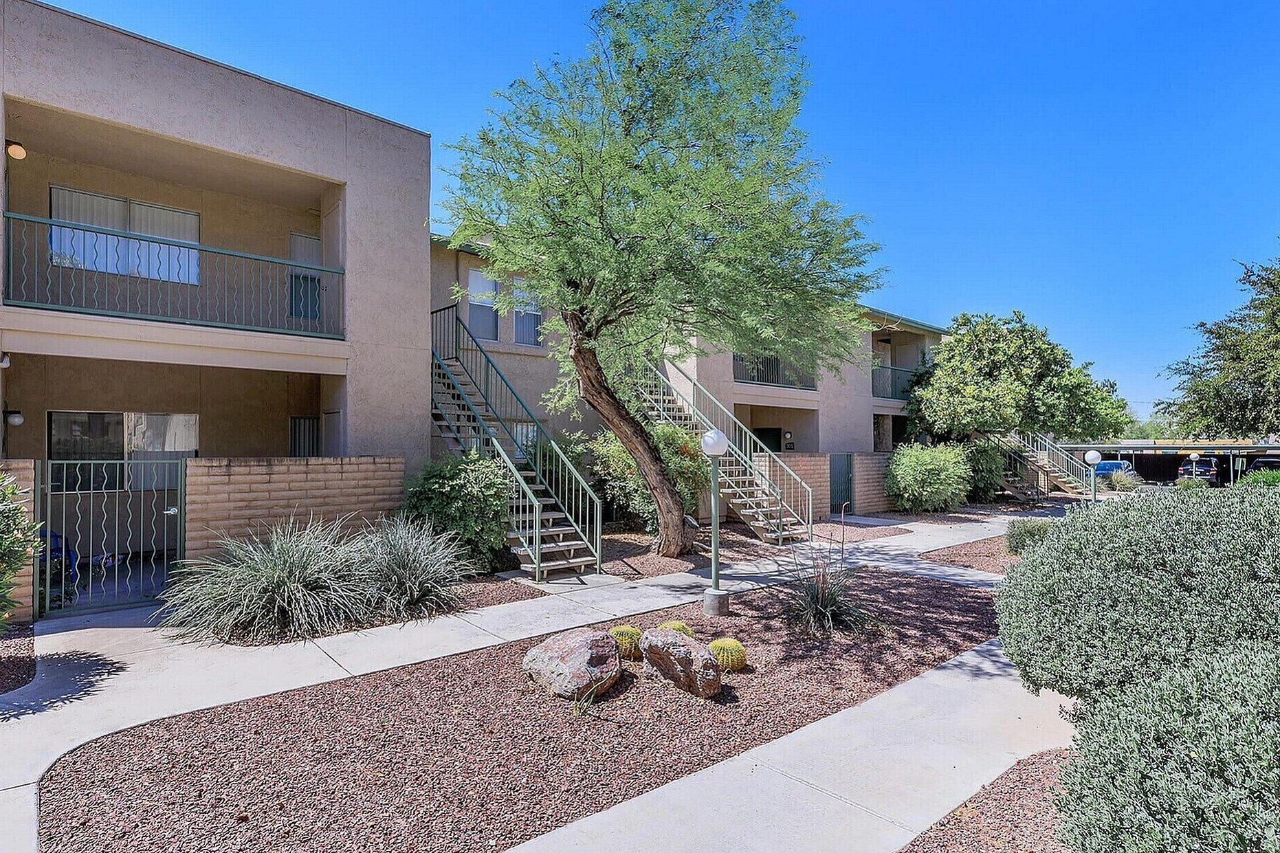 Charming apartment complex with beautiful landscaping and modern walkways, featuring balconies, lush greenery, and vibrant desert plants under a clear blue sky.