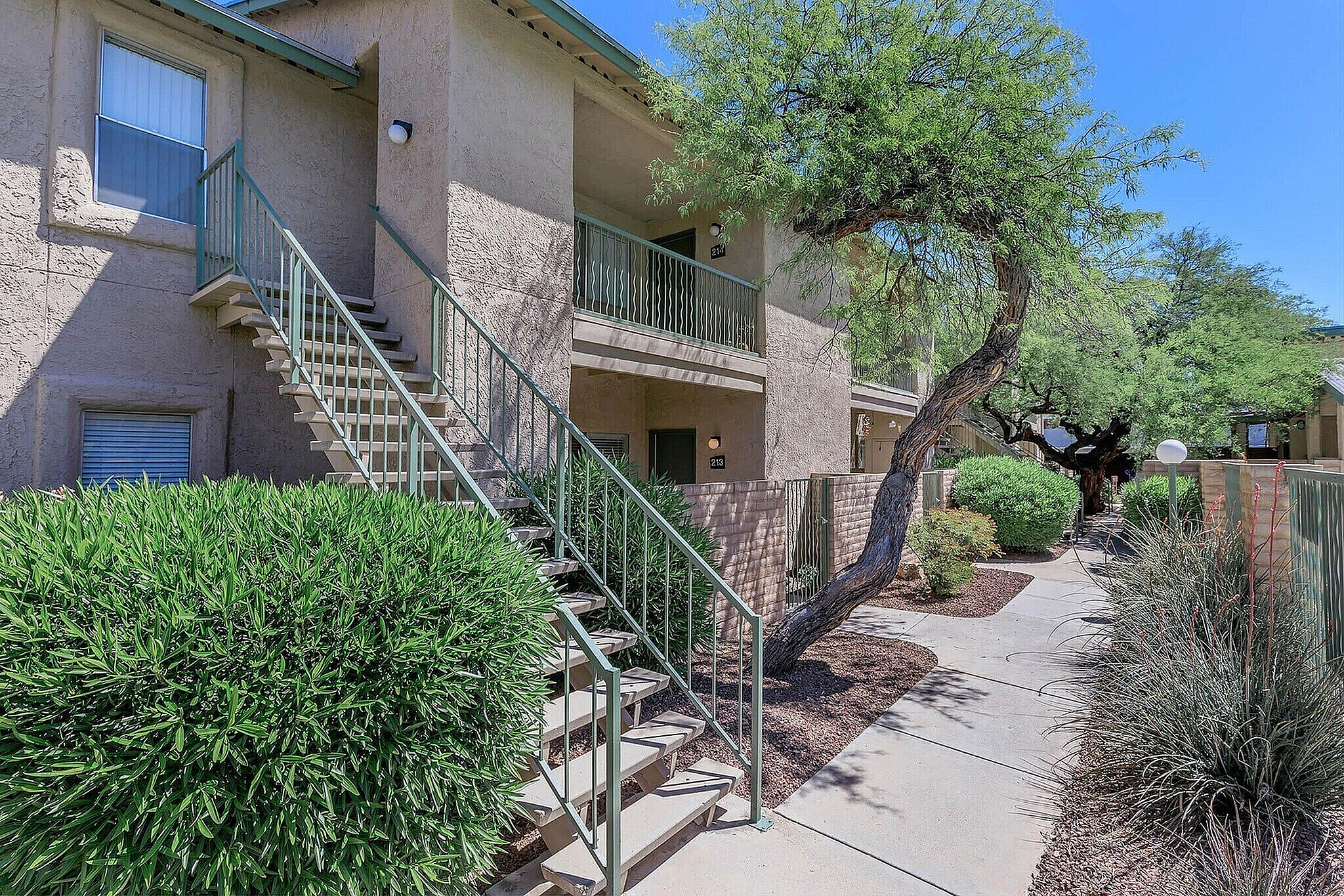 Inviting apartment exterior with lush landscaping, staircase leading to upper level, and vibrant greenery beneath a clear blue sky.