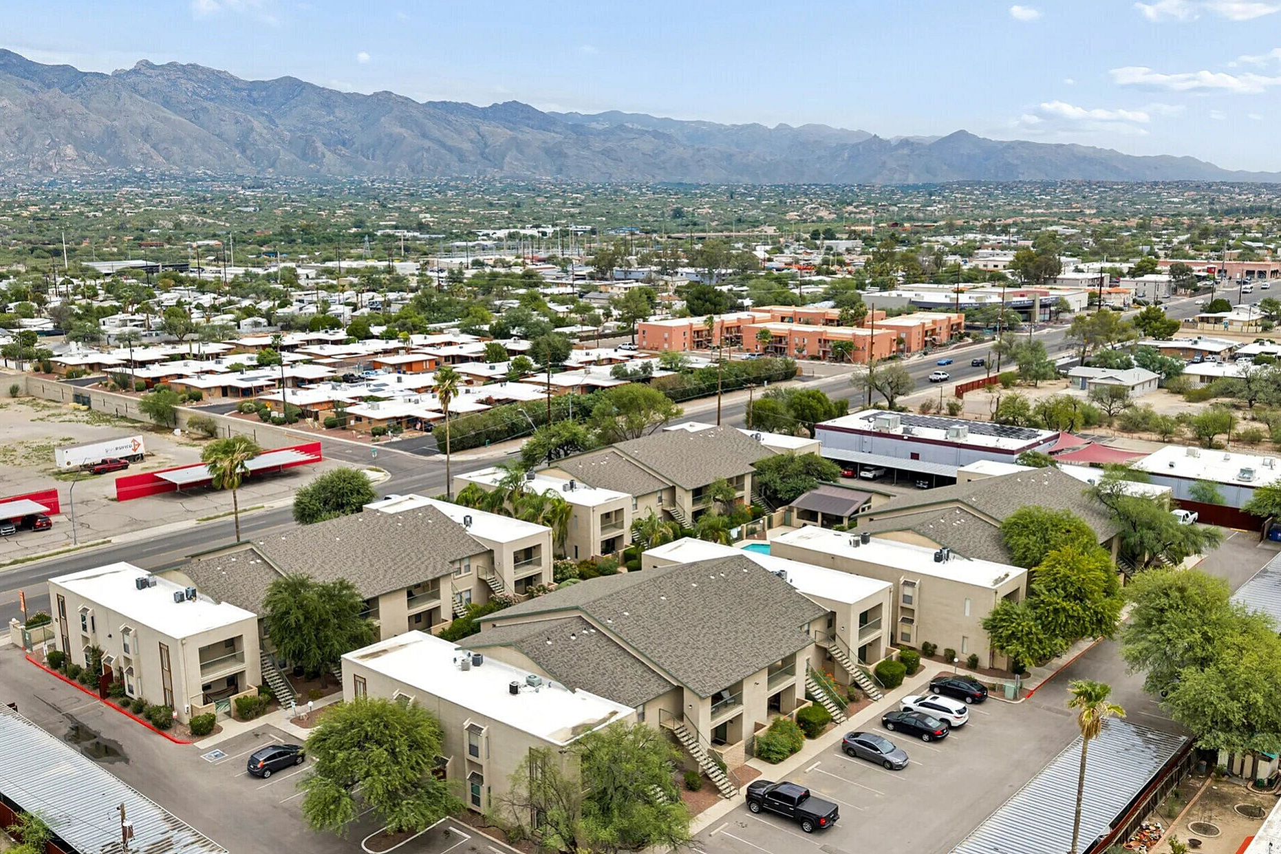 Aerial view of a modern apartment complex surrounded by lush greenery, stunning mountain backdrop, and nearby shopping area.