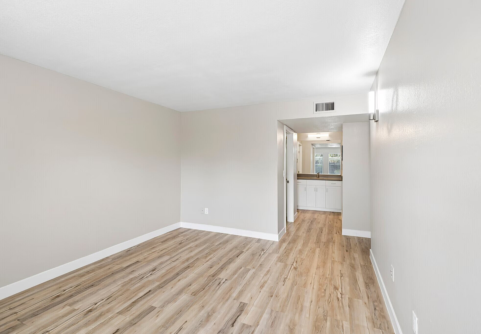 Bright apartment interior showcasing a spacious living area with sleek wooden flooring, neutral walls, and a well-lit kitchen entry.