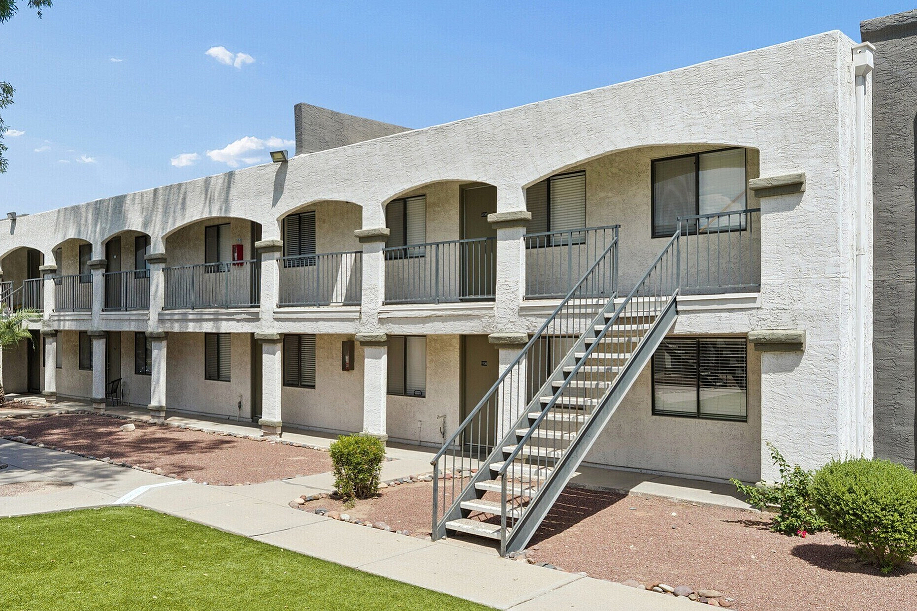 Charming two-story apartment building with arched balconies, surrounded by landscaped greenery and a welcoming walkway.