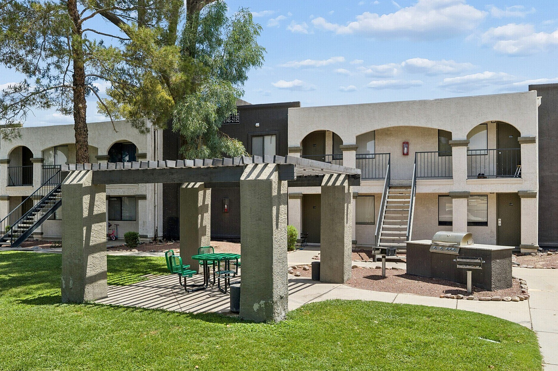 Charming apartment complex with a modern pergola and outdoor seating area, flanked by inviting greenery and a built-in barbecue grill.