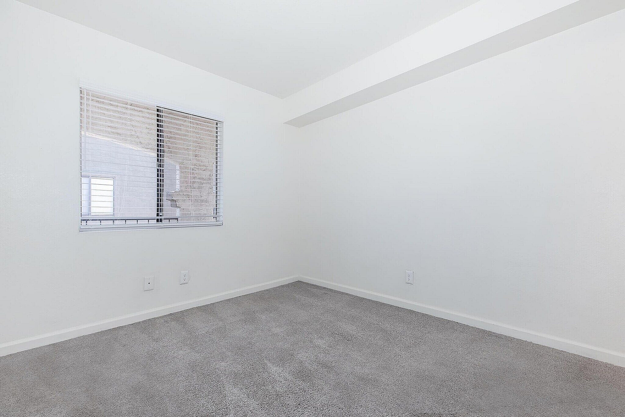 Bright, airy corner room featuring crisp white walls, plush carpet, and a large window with modern blinds, perfect for a serene living space.