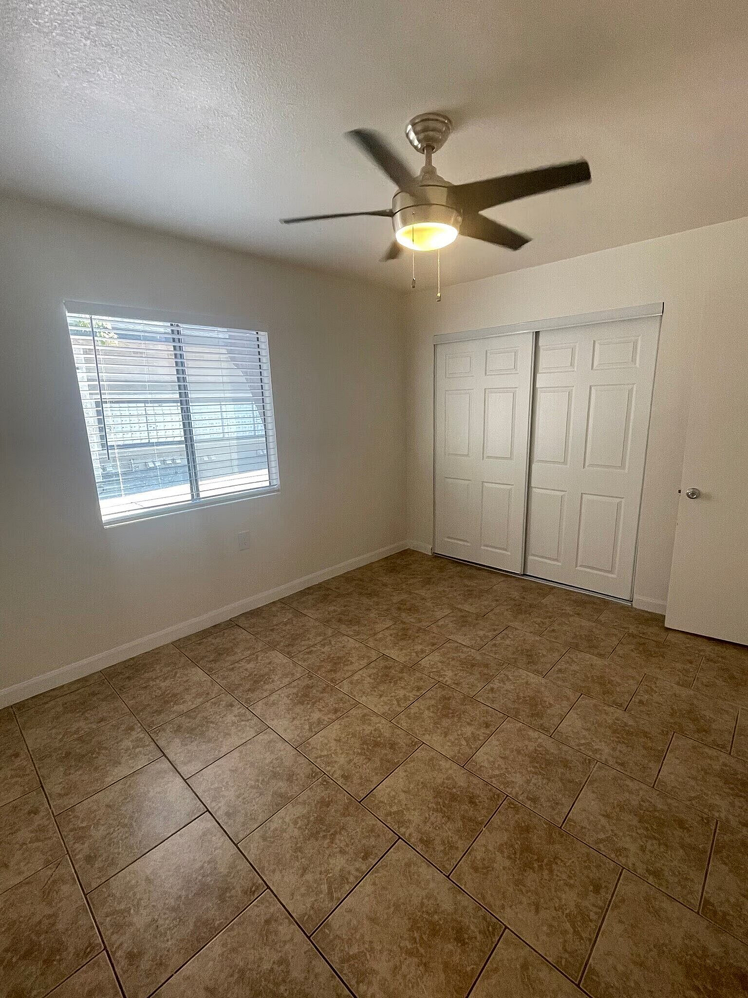 Bright and inviting room with modern ceiling fan, stylish tiled flooring, and ample closet space framed by fresh, natural light through a large window.