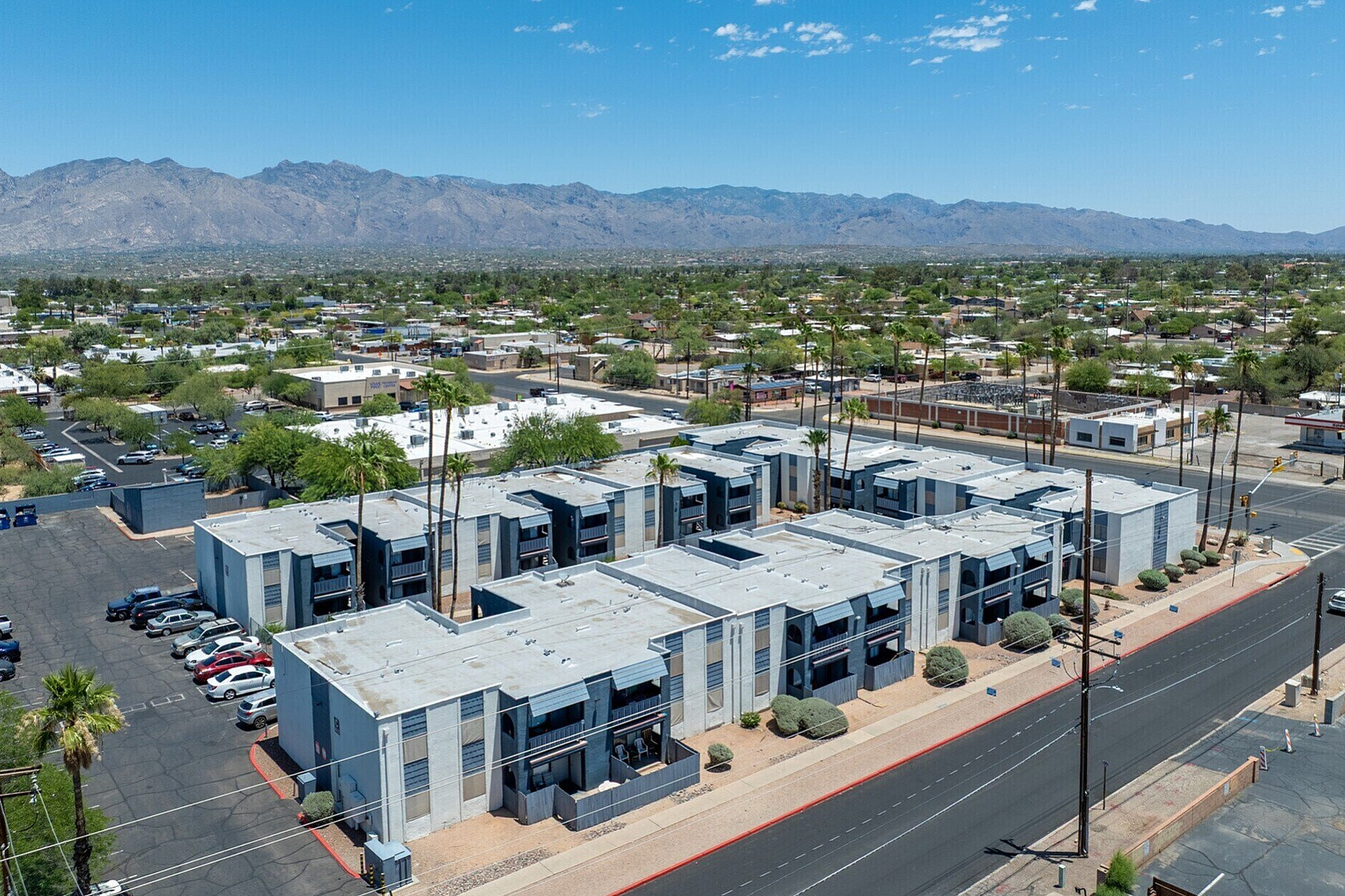 Aerial view of modern apartment complex with scenic mountain backdrop, lush greenery, and ample parking spaces beneath a clear blue sky.