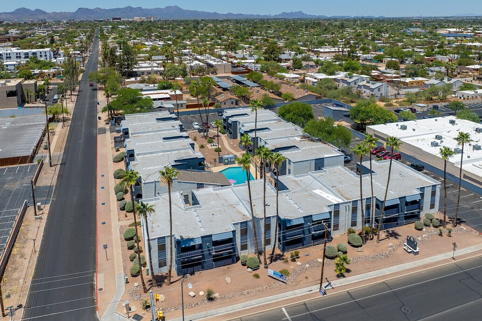 Aerial view of a modern apartment complex with a central swimming pool, surrounded by lush greenery and palm trees, showcasing a vibrant neighborhood and distant mountain views.