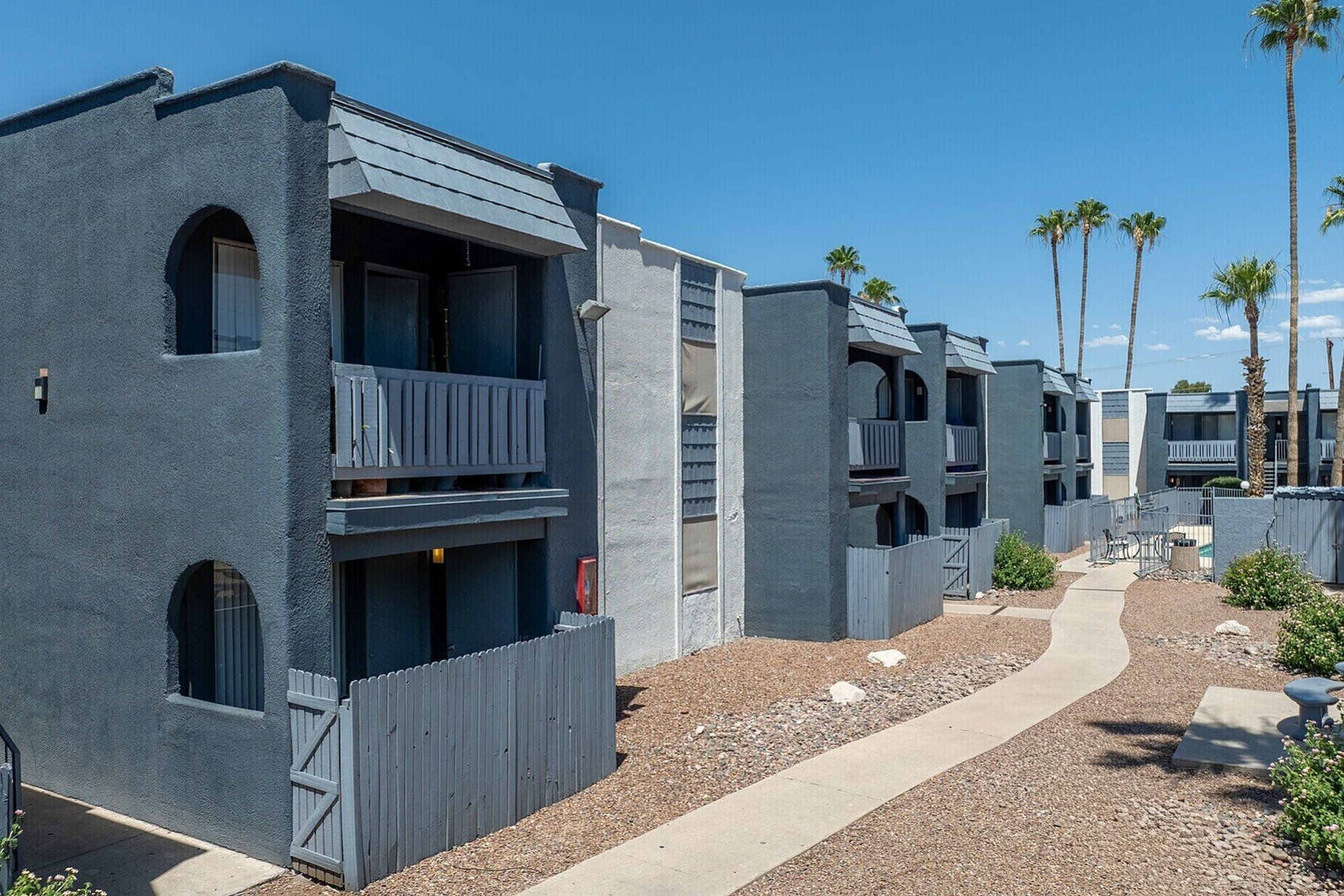 Modern apartment complex with unique architectural design, featuring private balconies and a serene garden path surrounded by palm trees.