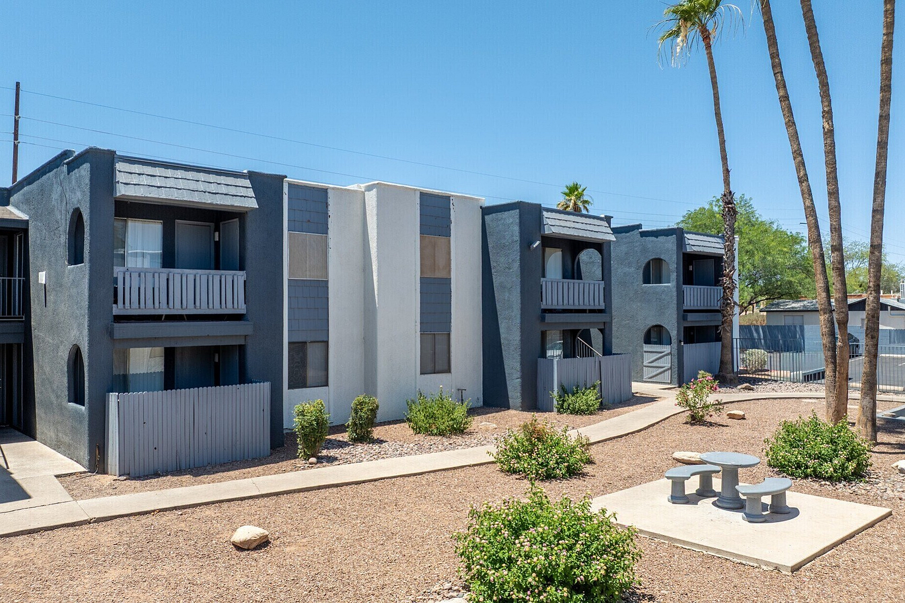 Charming apartment complex with modern dark blue and white exterior, featuring private balconies and beautifully landscaped gardens with palm trees.
