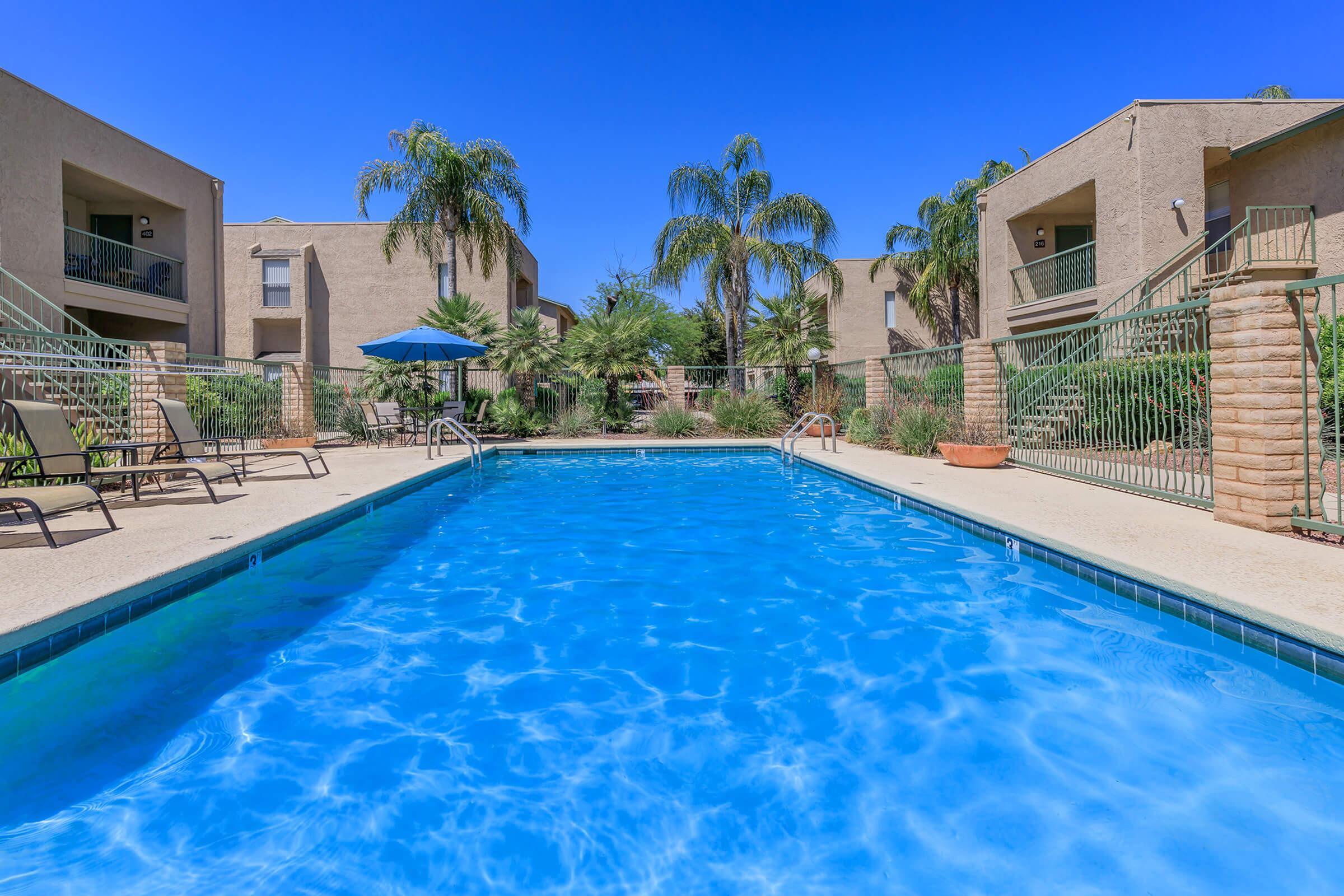 Inviting apartment complex featuring a sparkling blue swimming pool surrounded by palm trees and sun loungers under a vibrant clear sky.